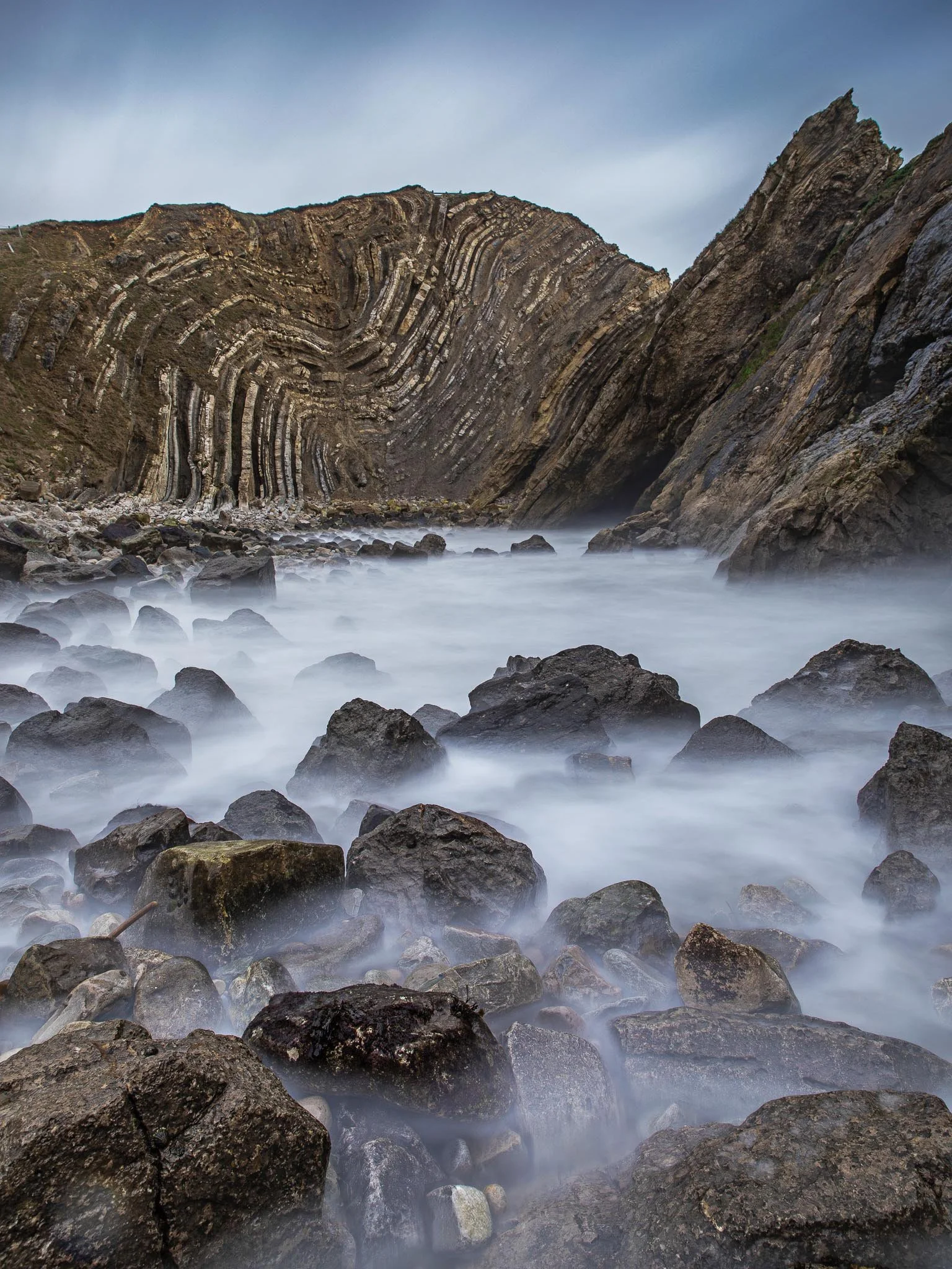Ocean waves crashing against rocks with a dramatic, layered cliff in the background under a cloudy sky.
