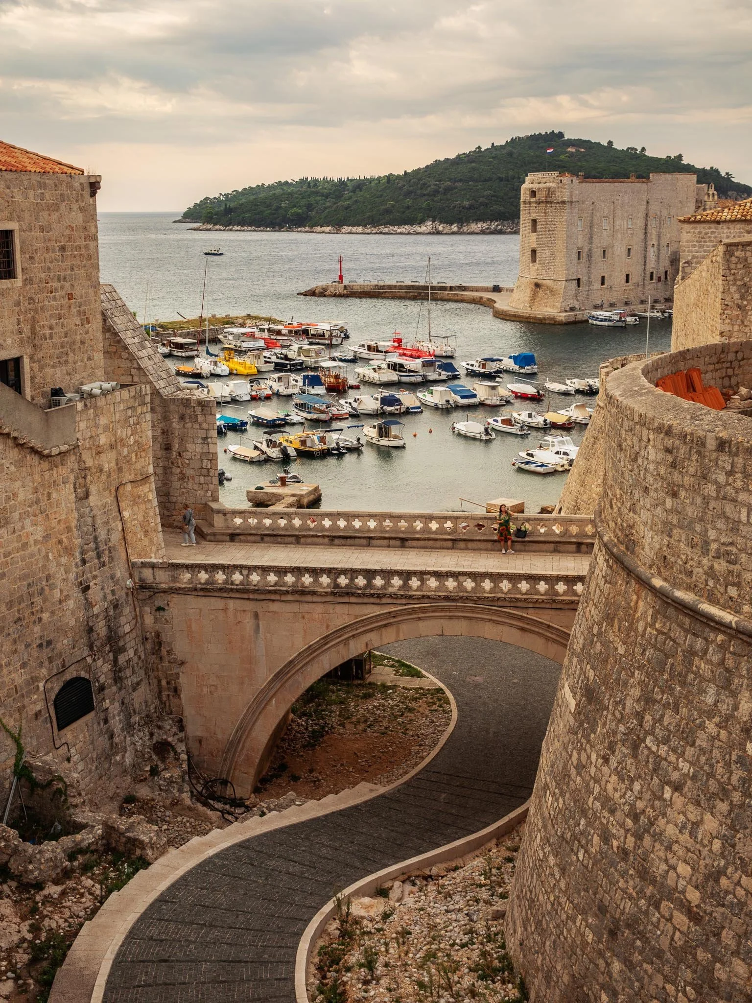 A harbor with numerous boats docked, surrounded by historic stone buildings and a stone fortress with a flag on top, overlooking a body of water with a forested hill in the background.