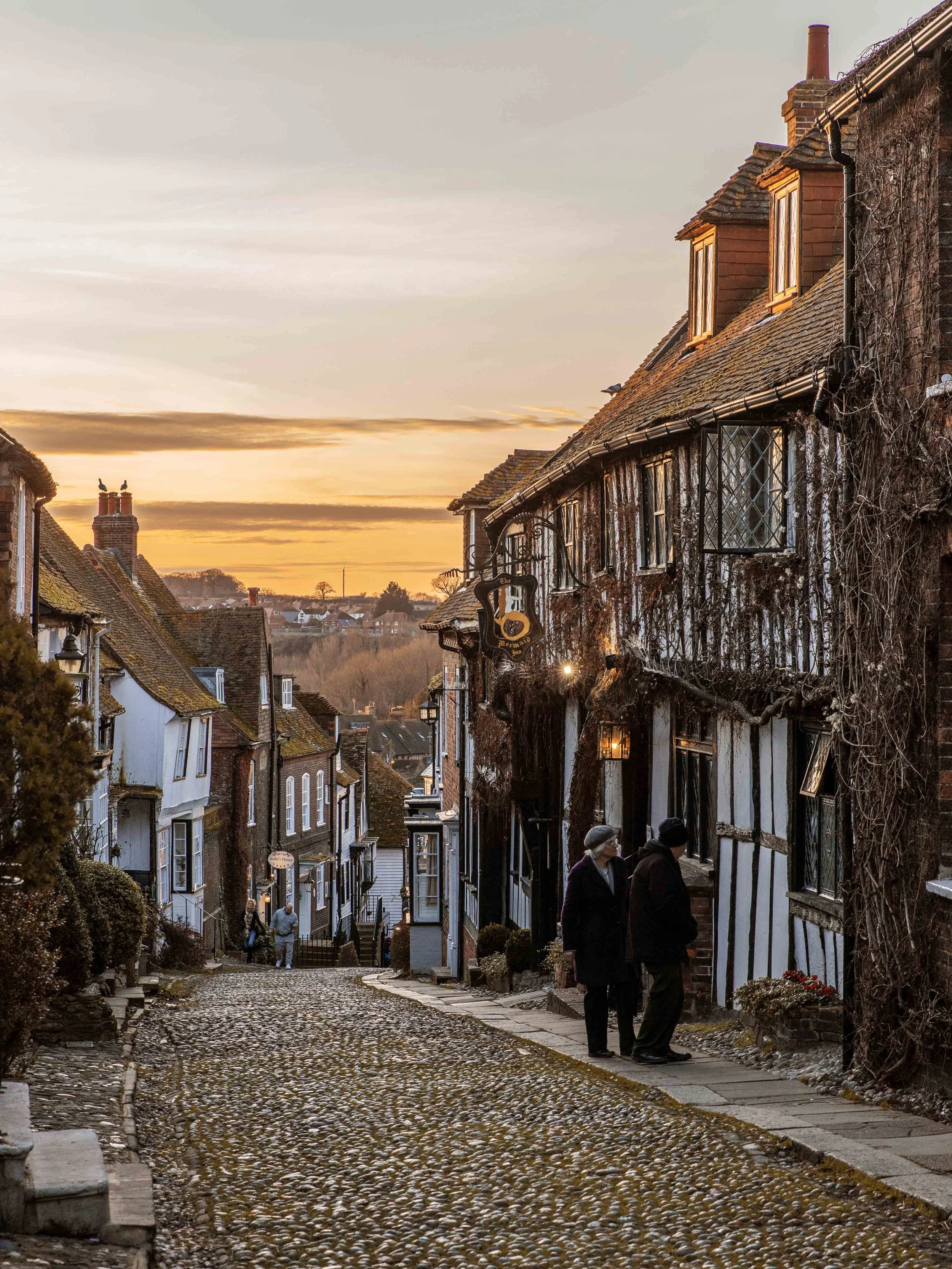 A cobblestone street in a historic town at sunset, lined with old brick and timber houses, with two people walking and talking in the foreground.