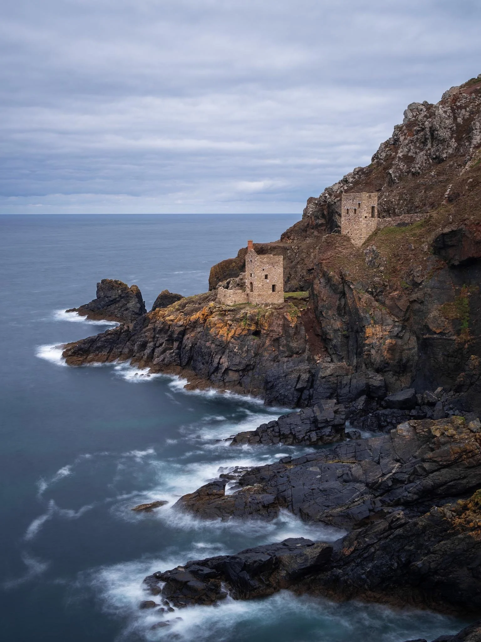Two stone ruins on a rocky cliff overlooking the ocean with cloudy sky.