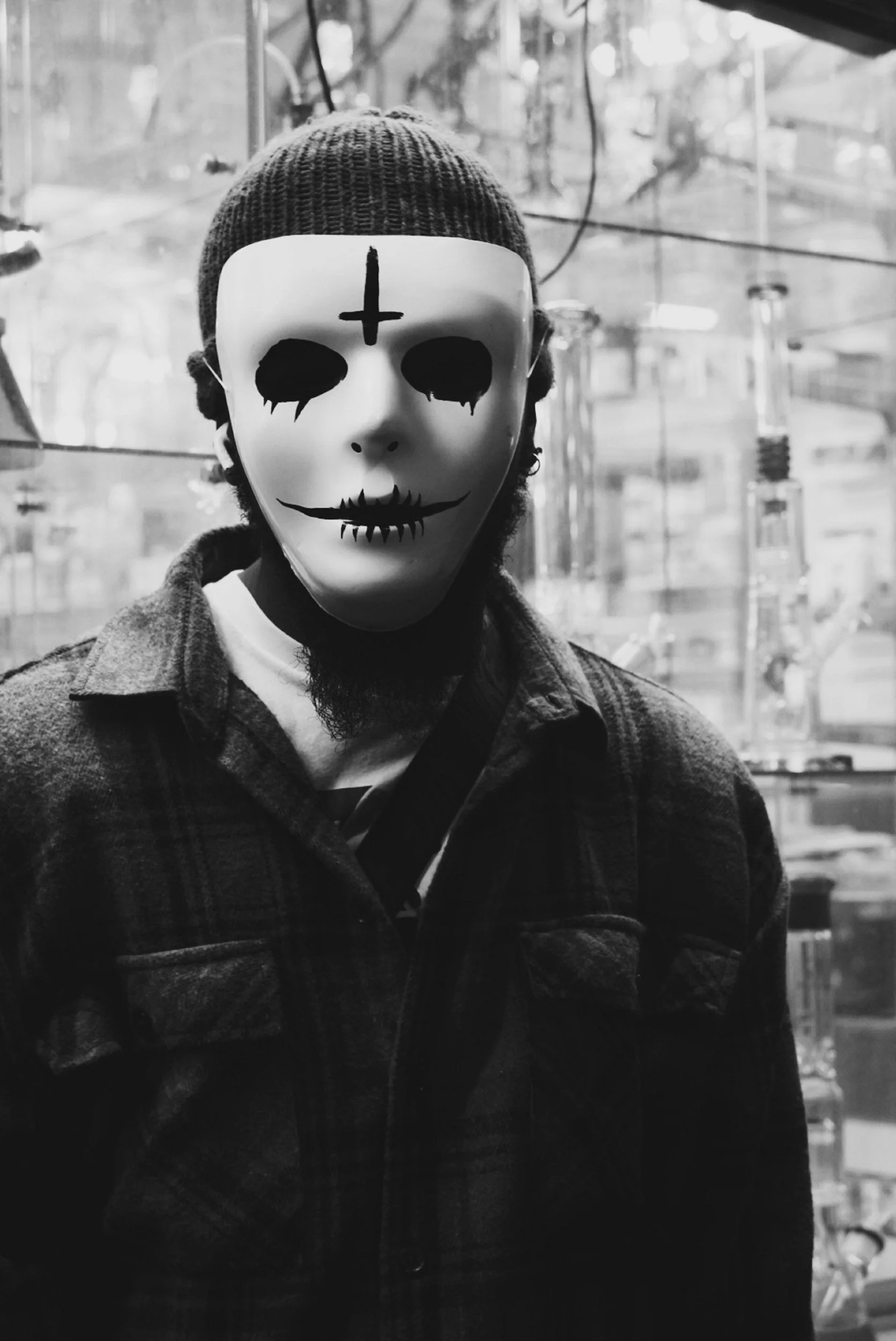 Person wearing a skeleton-shaped mask with black tears and stitched mouth design, a knit beanie, and a plaid jacket, standing indoors with a blurred glass display in the background.