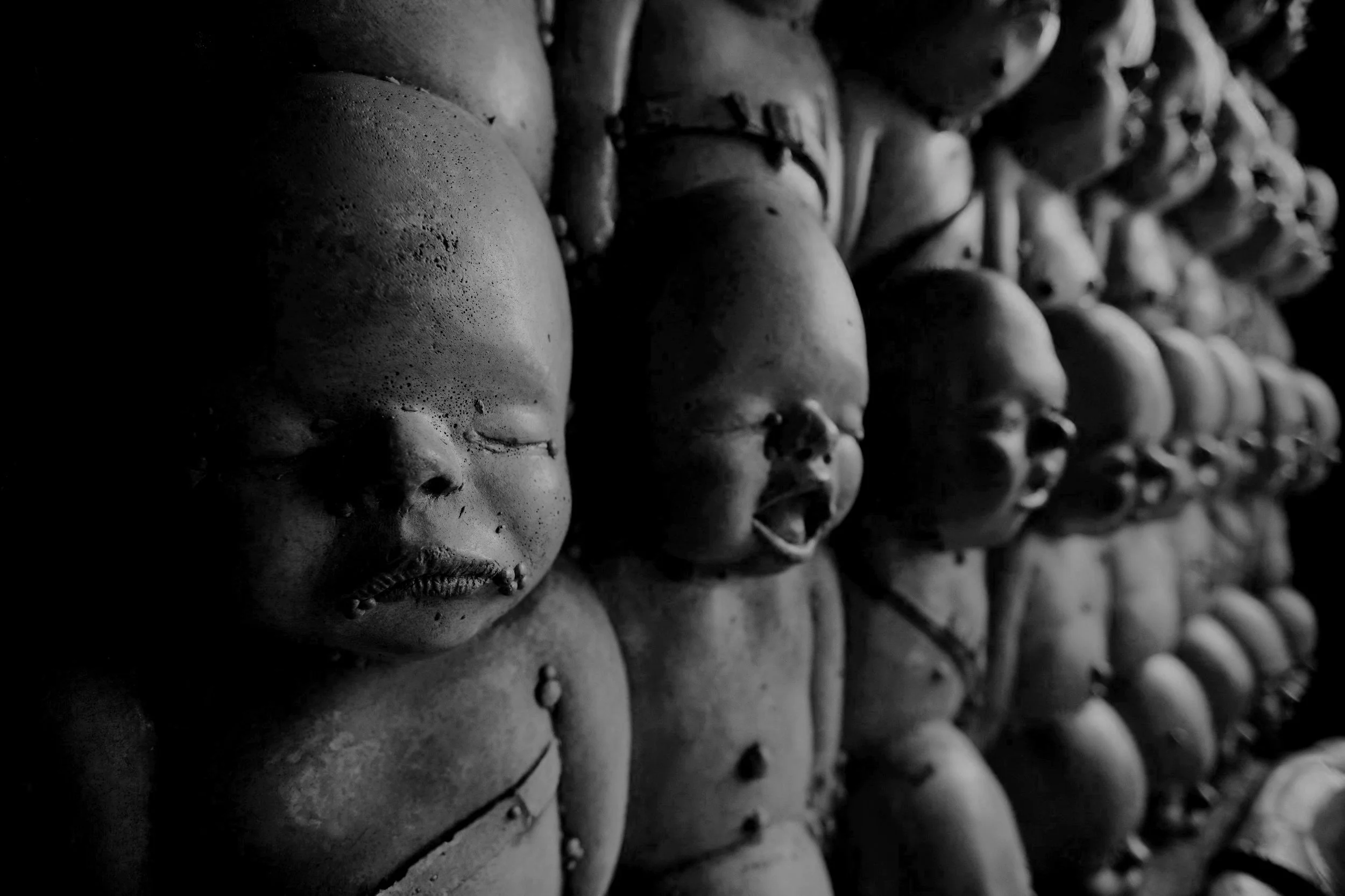 Row of clay or ceramic dolls with closed eyes and open mouths, arranged in a line, in black and white.
