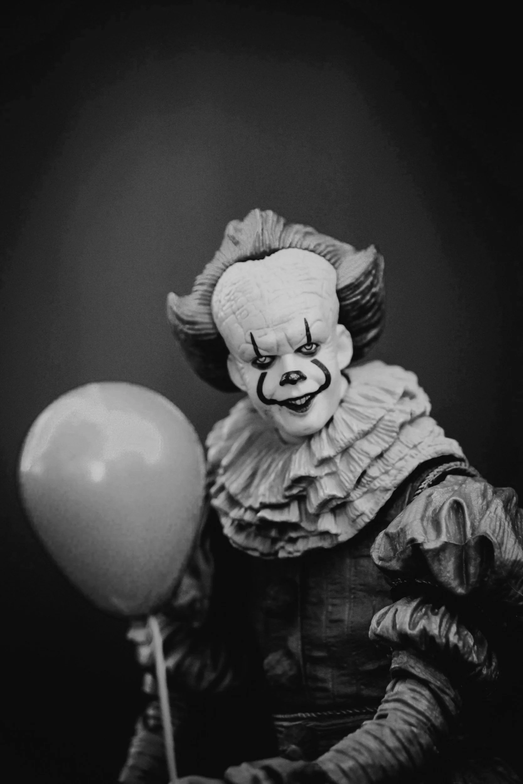 A creepy clown doll with exaggerated facial features, clown makeup, and curly hair, holding a balloon against a dark background.