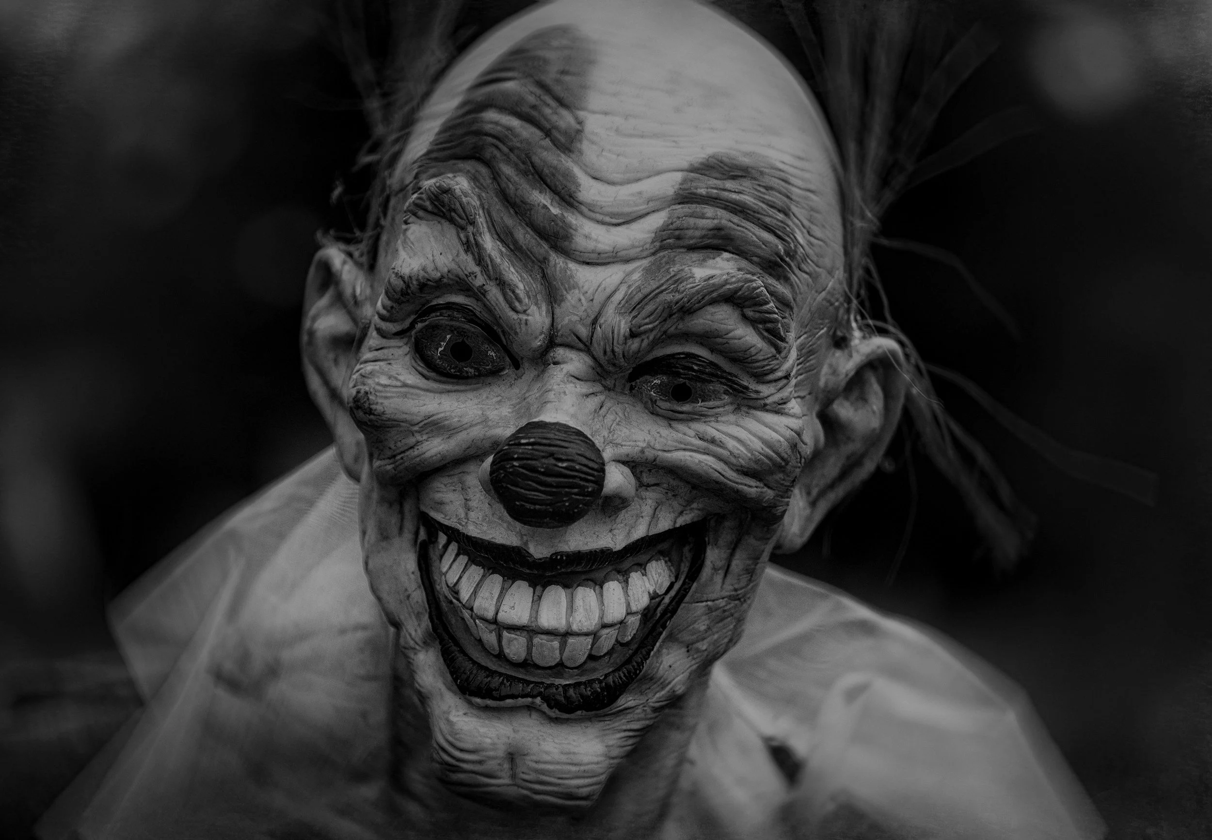 A close-up black and white photo of a creepy clown mask with exaggerated wrinkles, a big nose, and a wide, toothy grin.