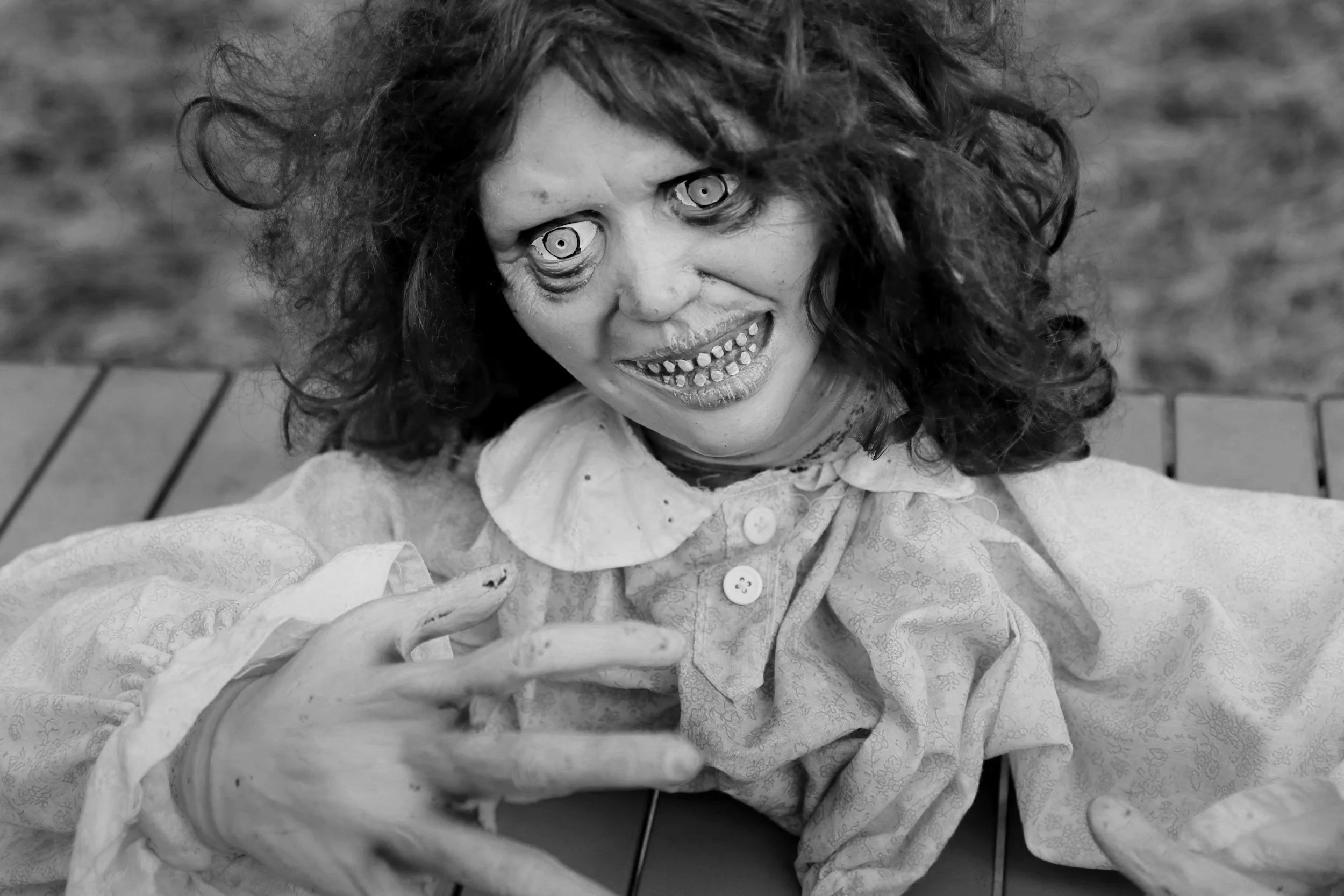 A creepy, zombie-like woman with curly hair and exaggerated facial features, including large, bulging eyes, a wide open mouth with crooked teeth, and decayed skin, sitting on a bench outdoors wearing old fashioned clothing.