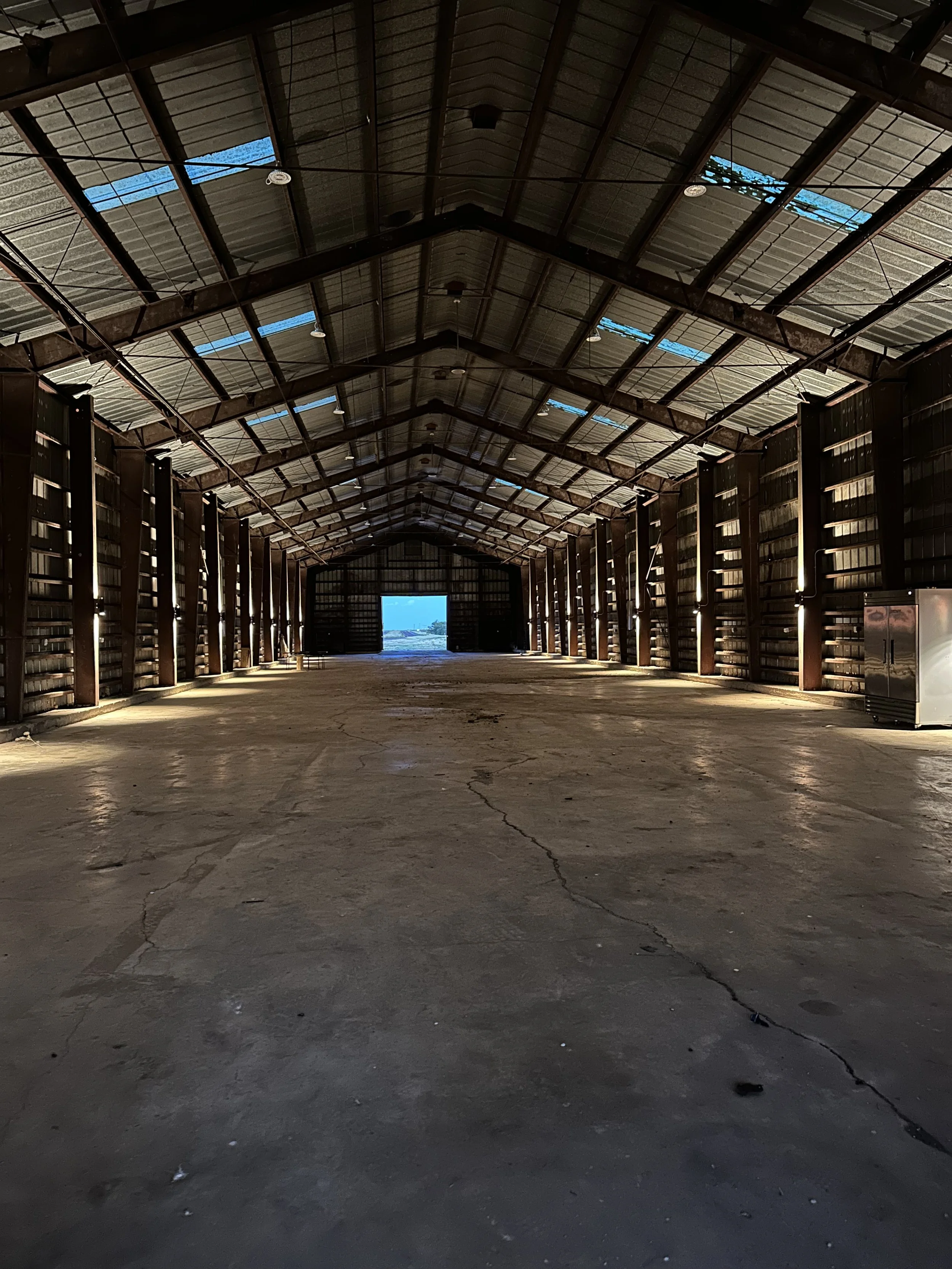 Inside view of a large, empty barn with metal walls and roof, sunlight streaming through small windows, concrete floor, and double doors open to outside.
