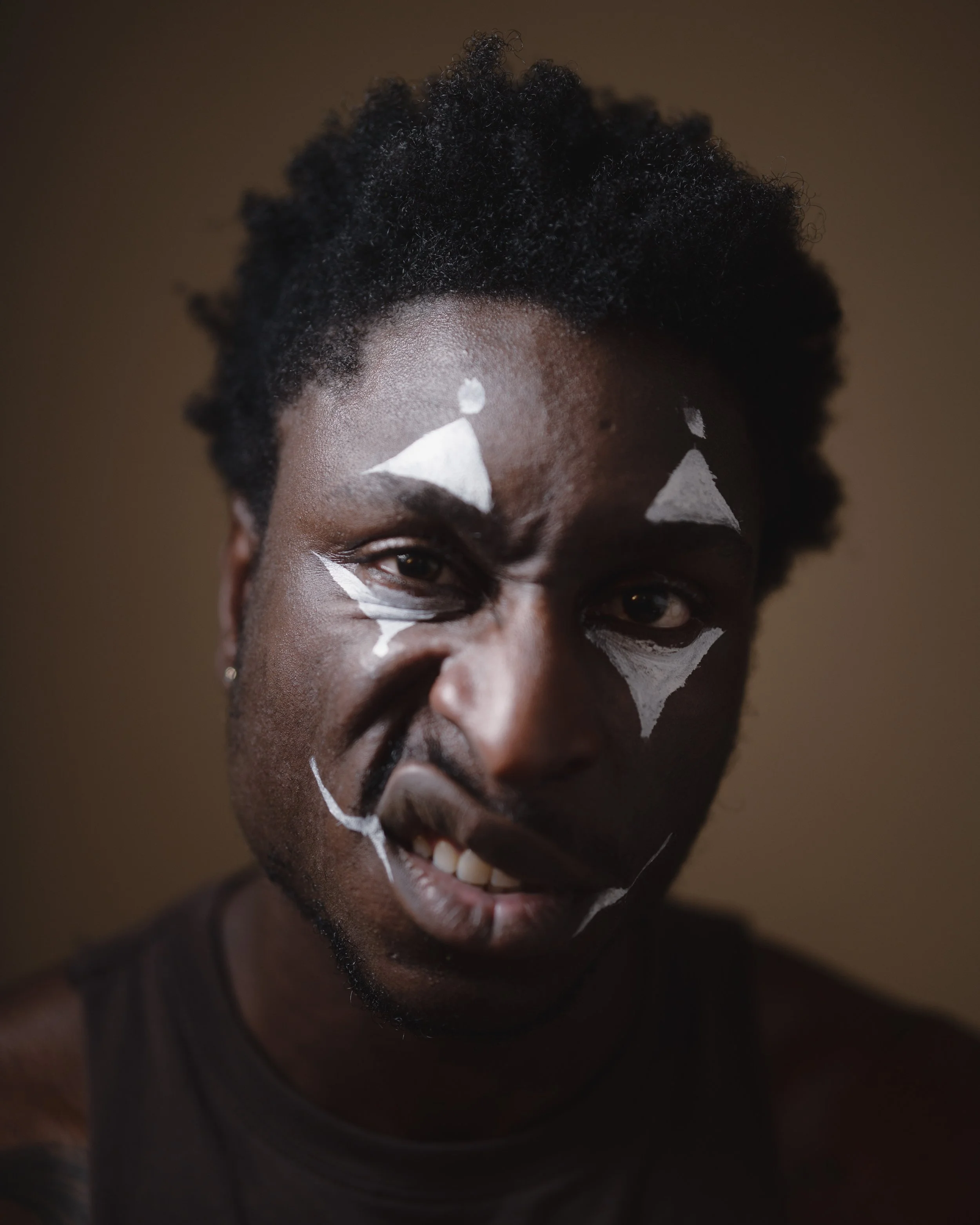 A person with dark skin and short, curly black hair has white paint or makeup on their face, including triangles above and below the eyes and lines on the cheeks.
