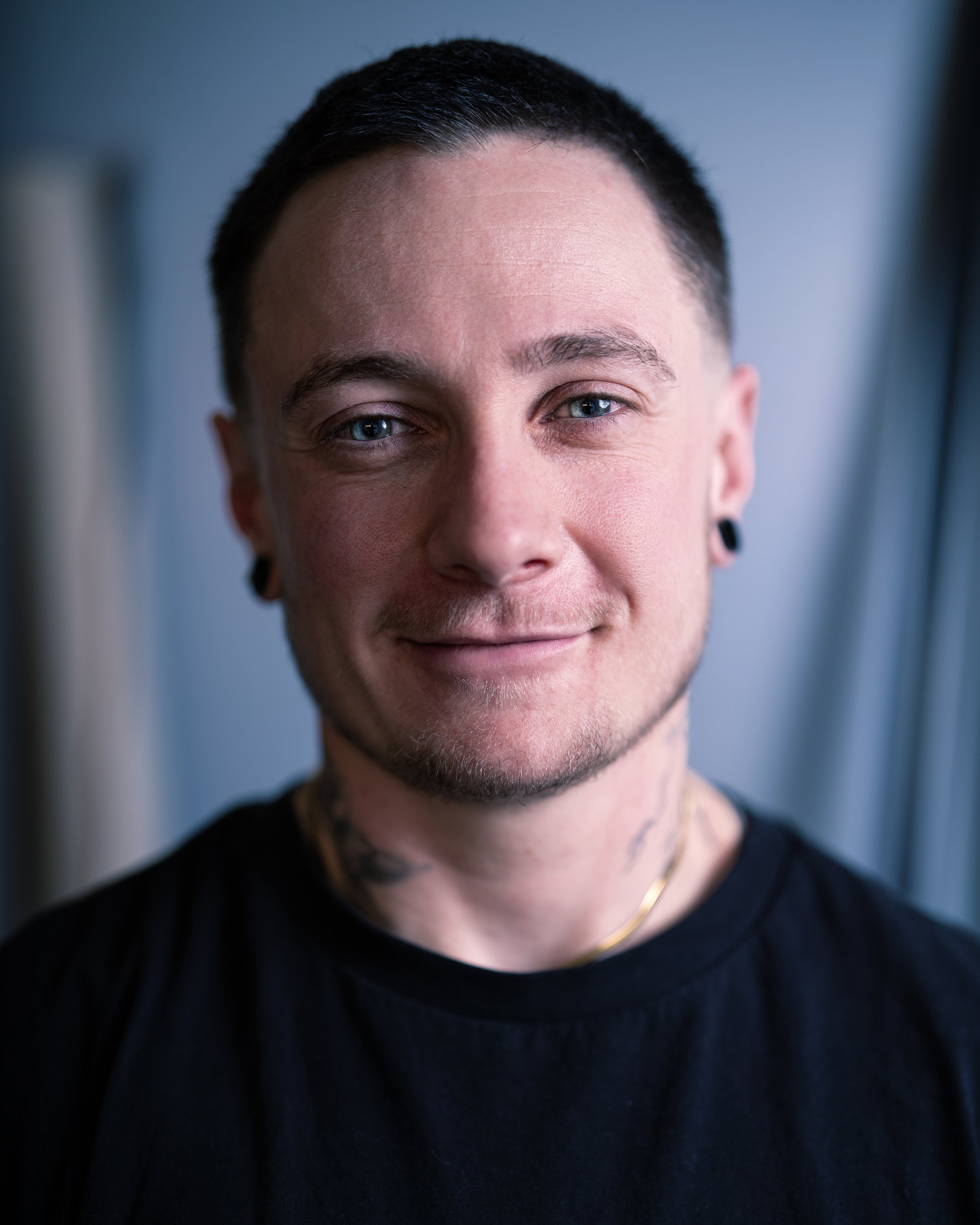 Headshot of a man with a buzz cut and blue eyes in front of a blue background wearing a black t-shirt