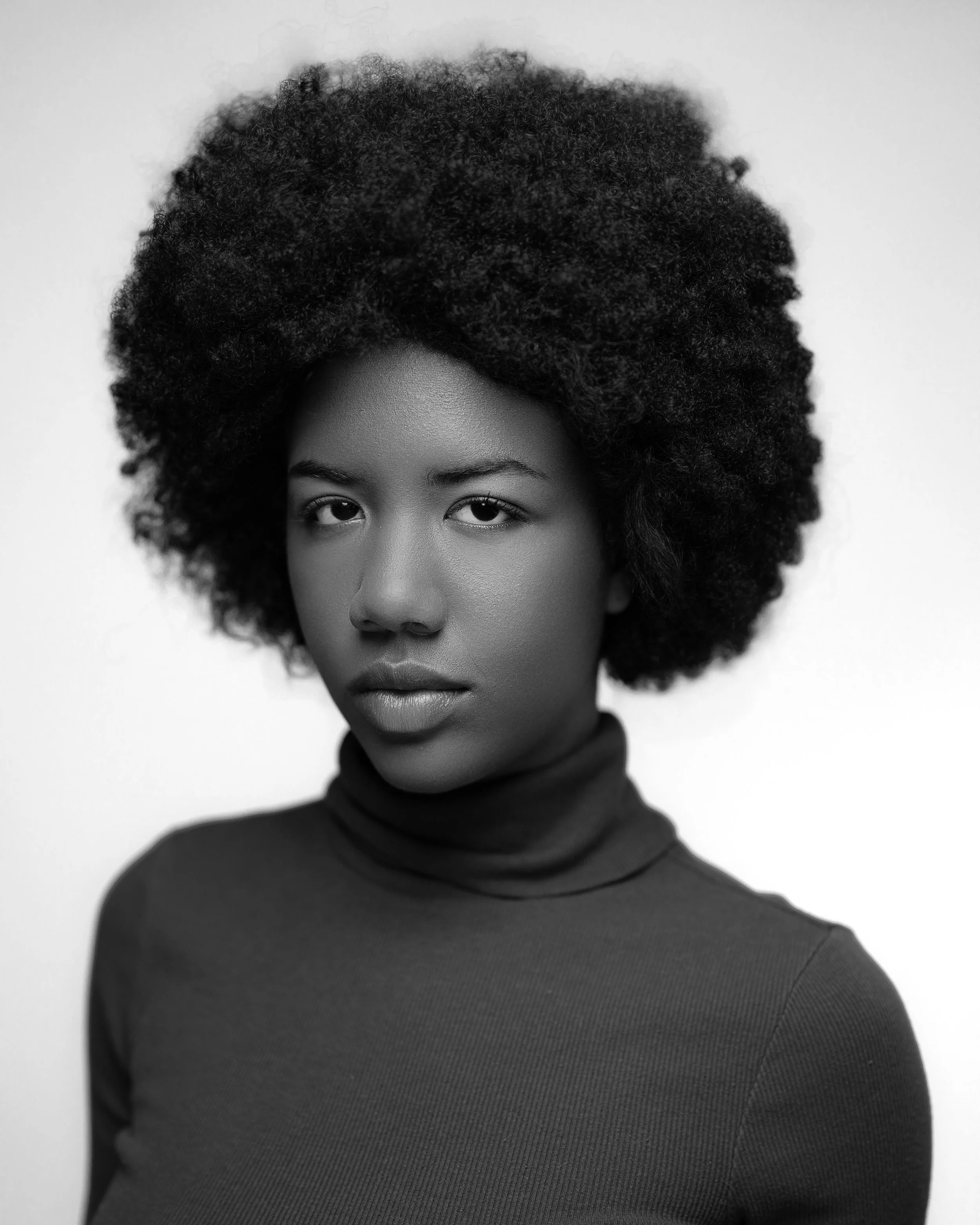 Close-up of a woman with dark skin and voluminous curly afro hair, wearing a turtleneck sweater, in black and white.