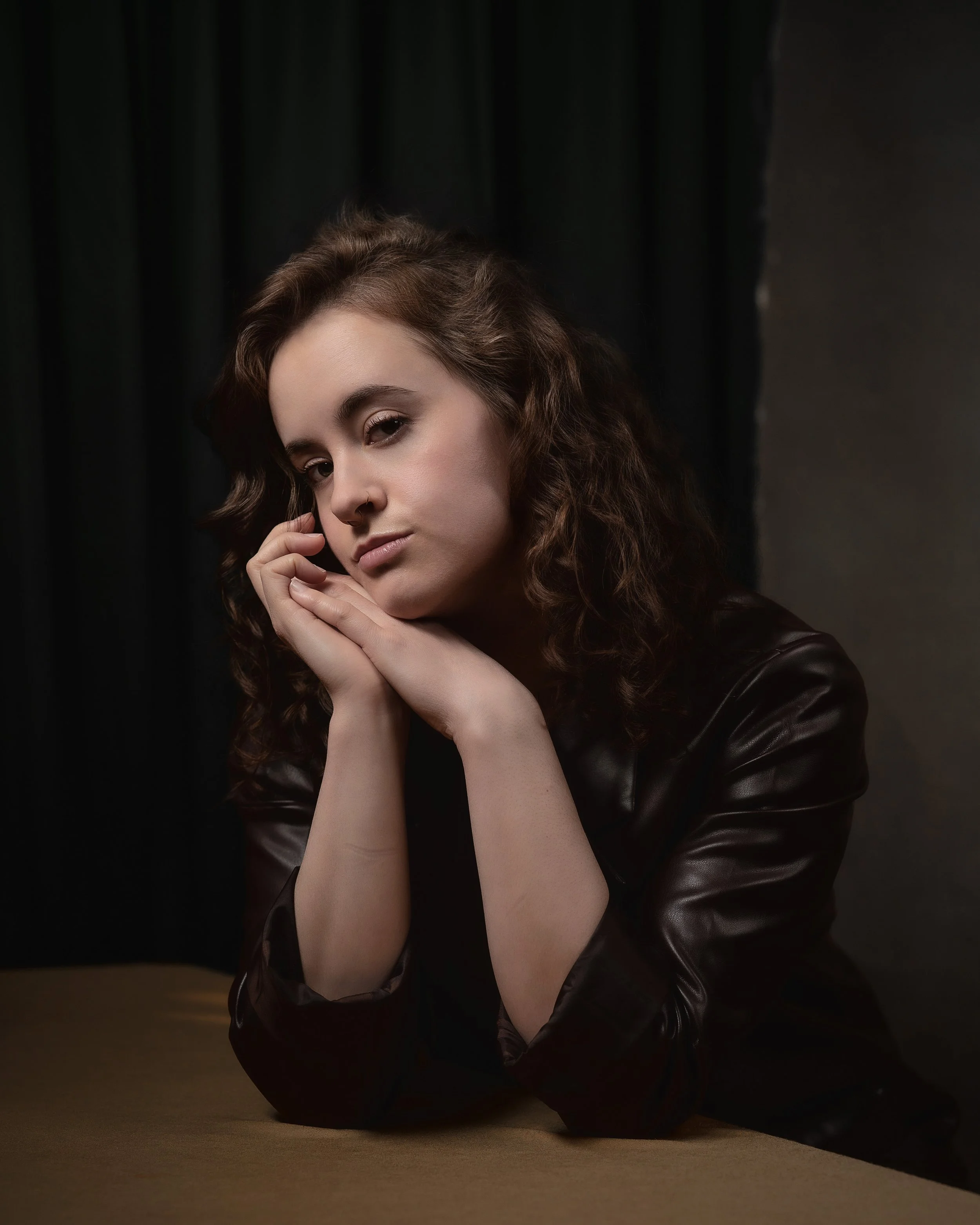 A woman with brown, curly hair and a nose ring is lying on the ground with her head resting on her hands. She is wearing a dark leather jacket and looks directly at the camera against a dark background.
