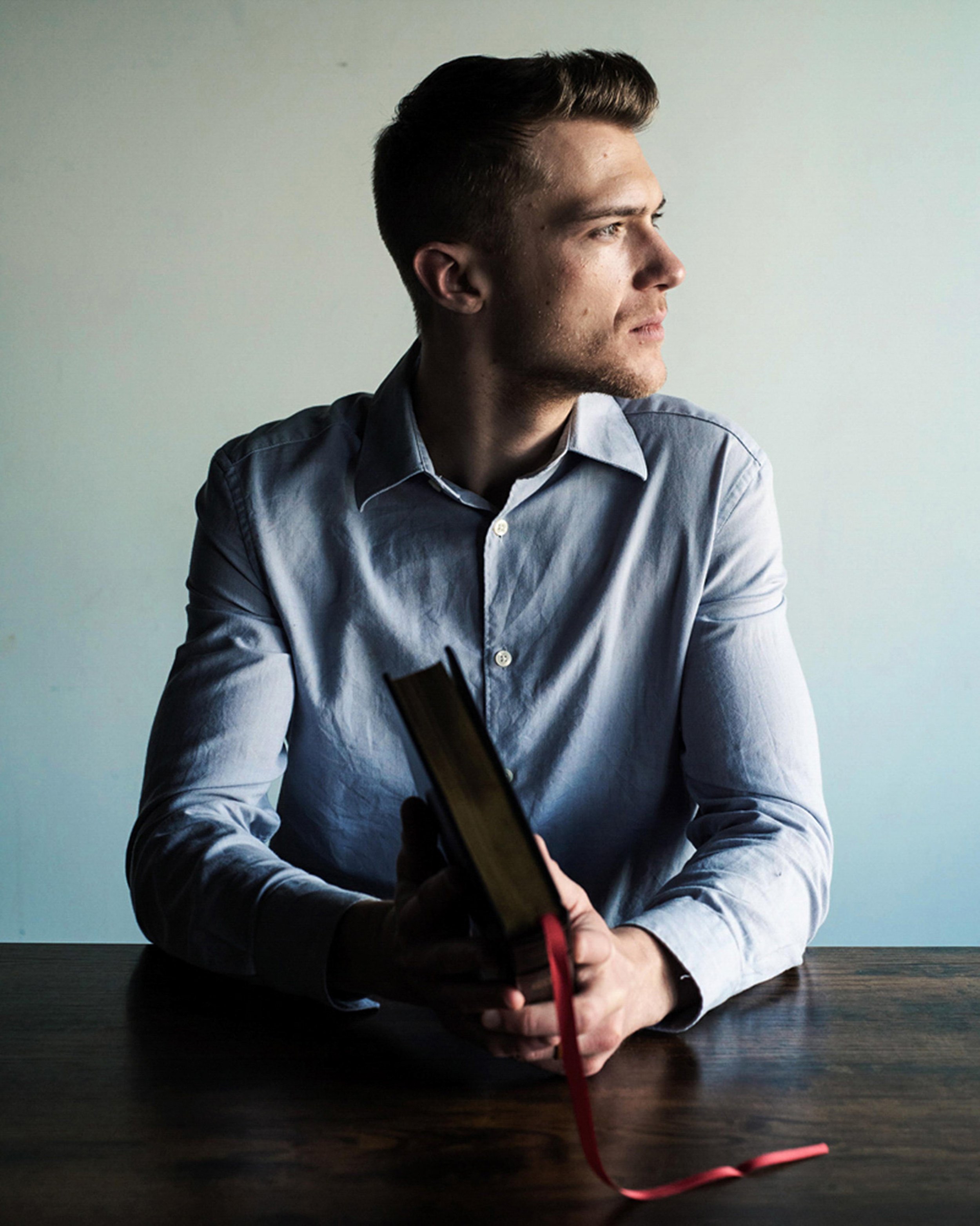 A young man with dark hair and a beard, wearing a light blue dress shirt, sitting at a dark wooden table, looking thoughtfully to the right, with a plain light-colored wall in the background.