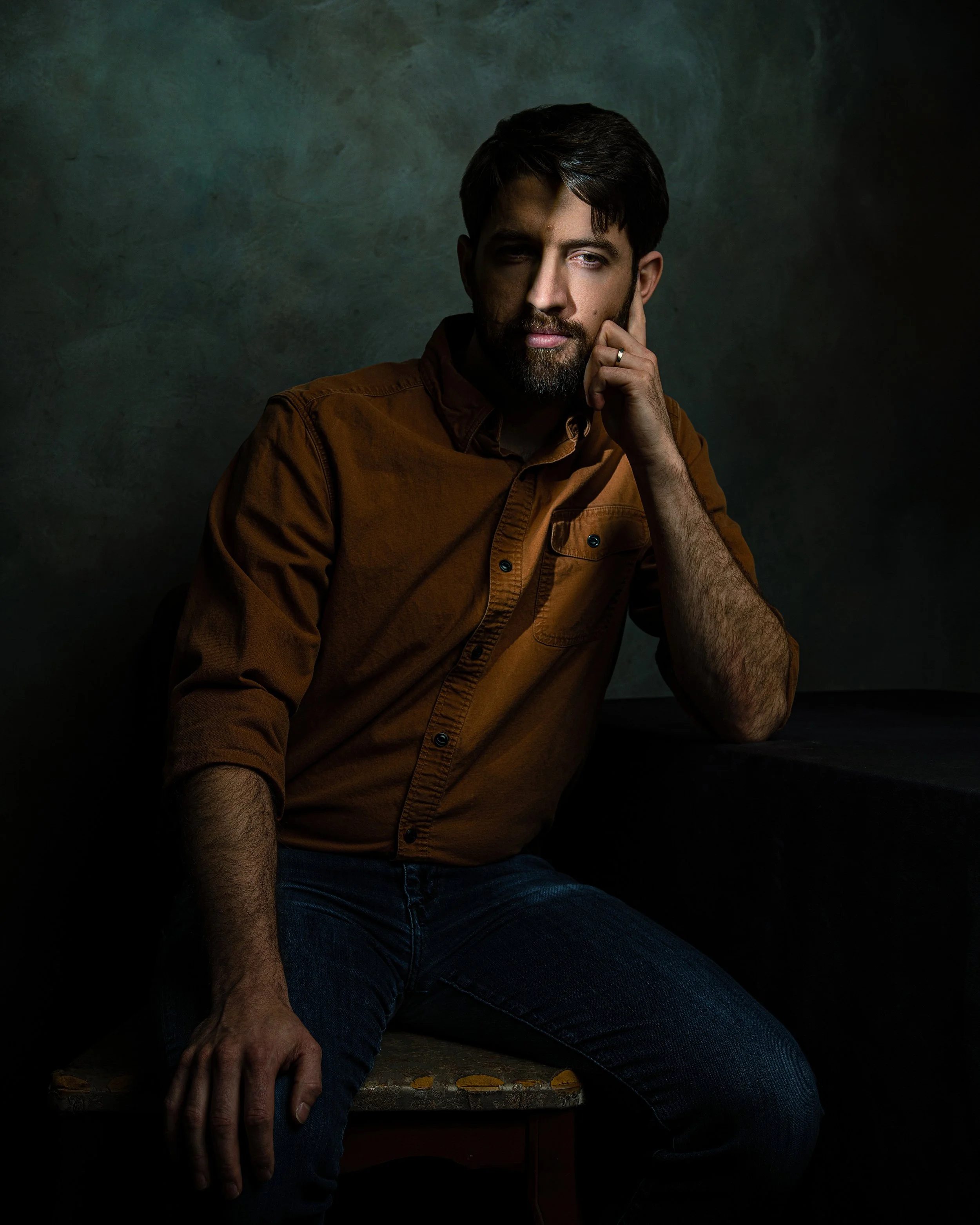 A man with dark hair, beard, wearing a brown shirt and blue jeans, sitting on a wooden bench with graffiti, in a dark setting, resting his head on his hand with a contemplative expression.