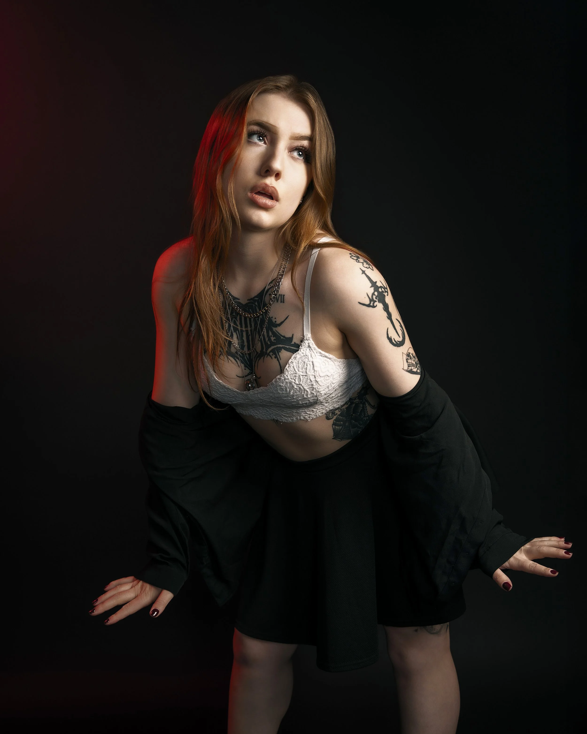 A young woman with long red hair and multiple tattoos poses against a dark background. She is wearing a white lace top and black pants, with her hands resting on the ground, fingers spread out, and nails painted dark red.