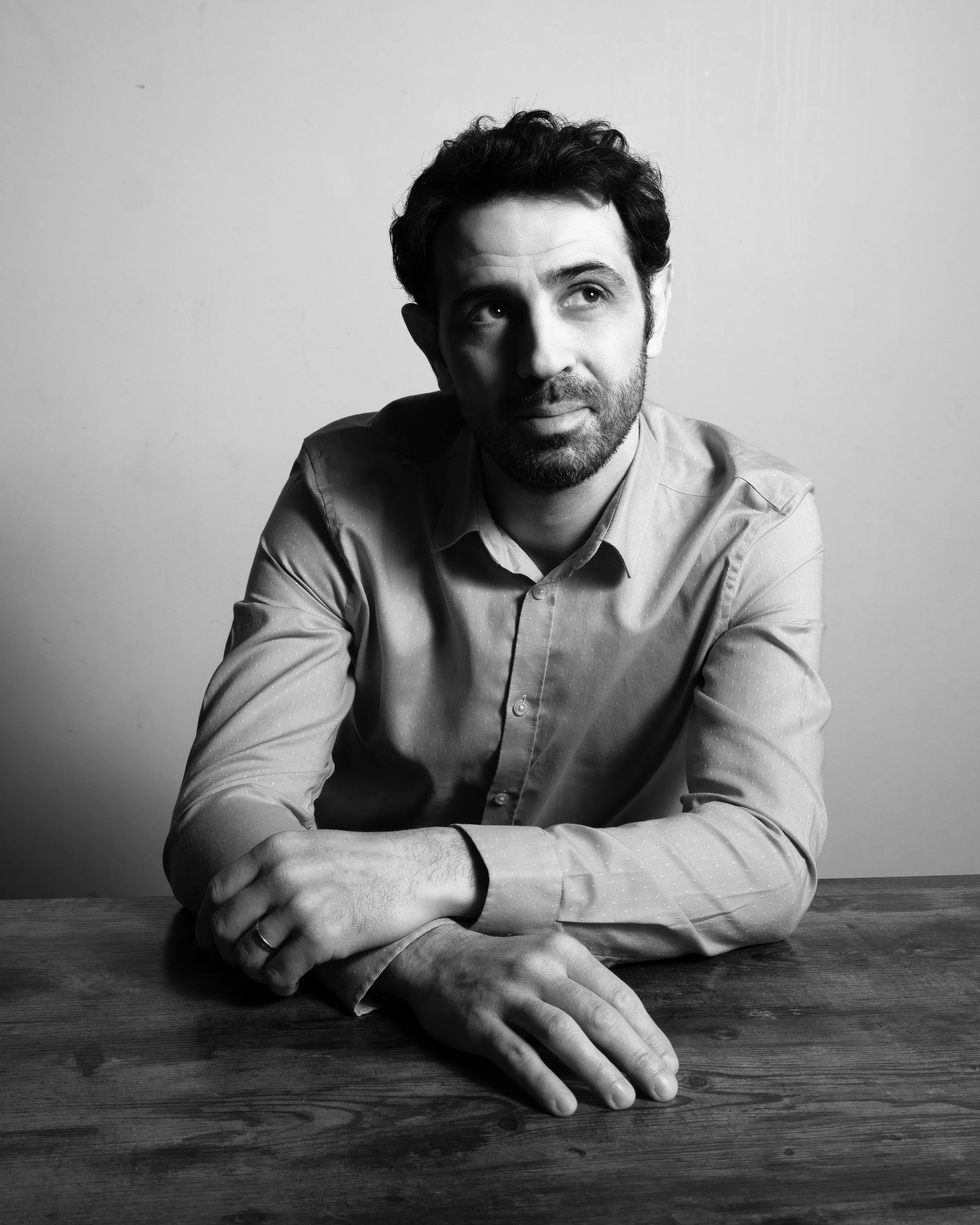 A black and white photo of a man sitting at a wooden table, with his left arm across it. He is wearing a collared shirt, has dark hair and a beard, and is looking slightly upward and to his left.