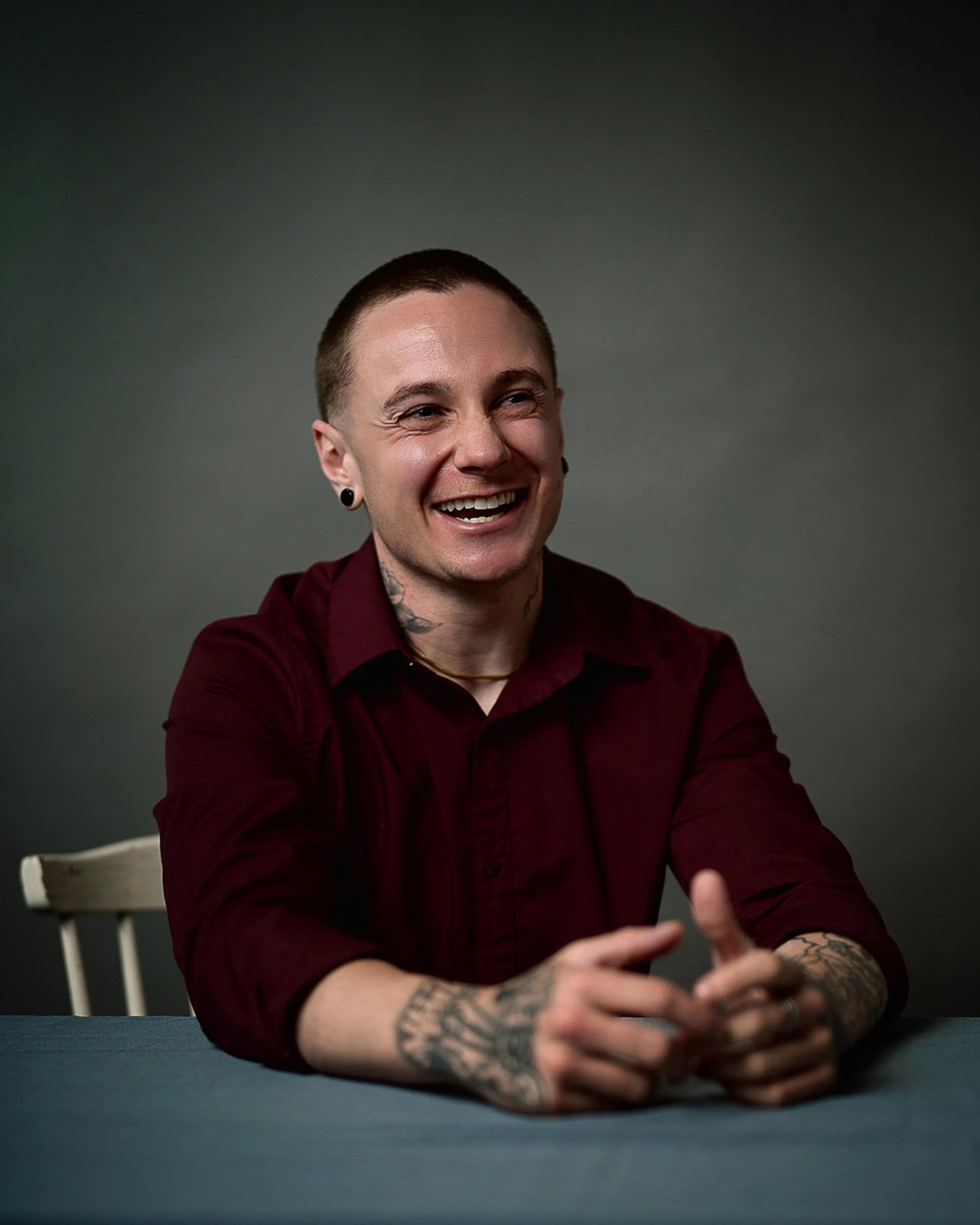 A young man with short hair and tattoos on his arms, wearing a maroon shirt, smiling and laughing while sitting at a table with his hands clasped.