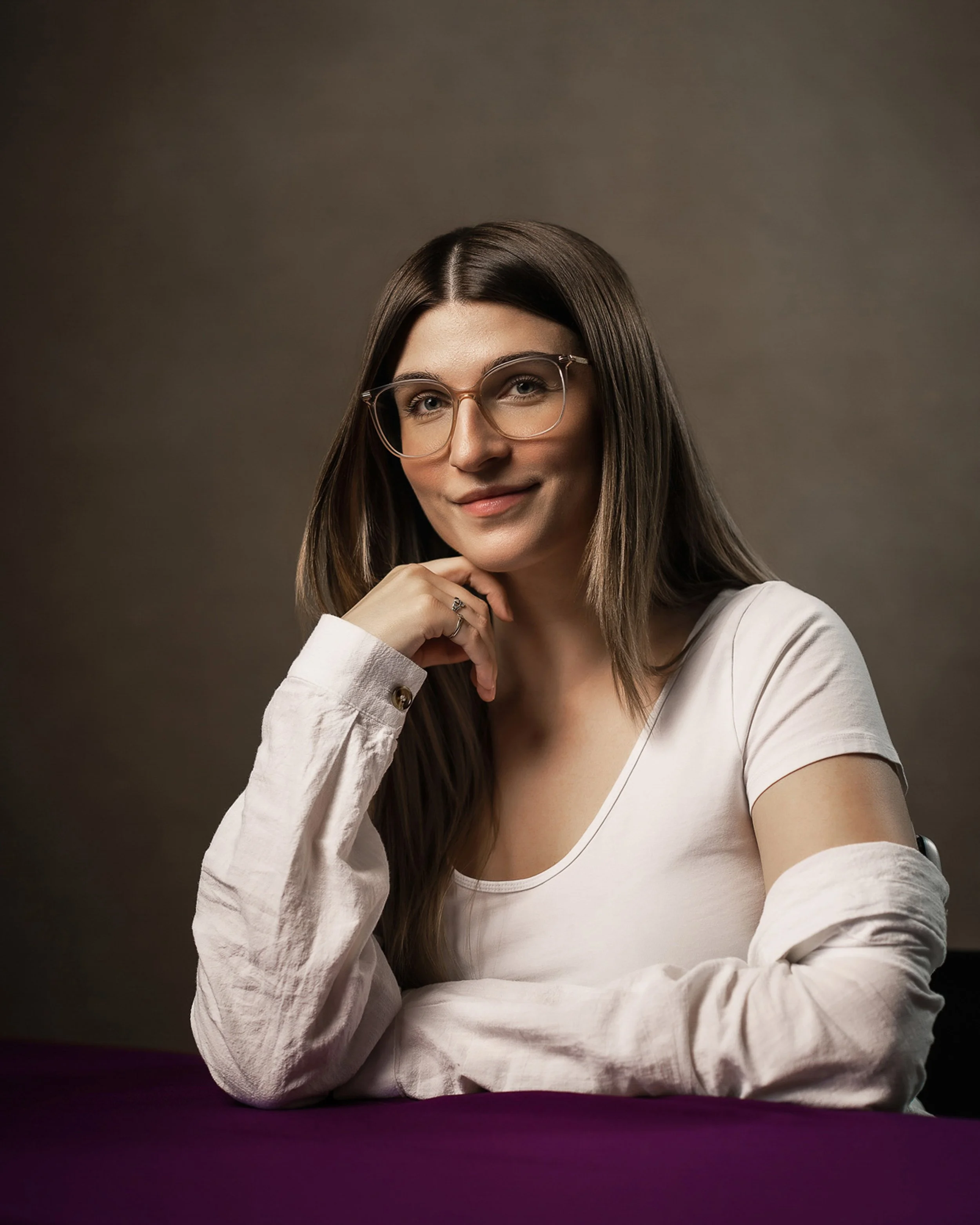 A woman with long brown hair wearing glasses with clear frames, smiling and resting her chin on her left hand, dressed in a white long-sleeve shirt, against a plain brown background.