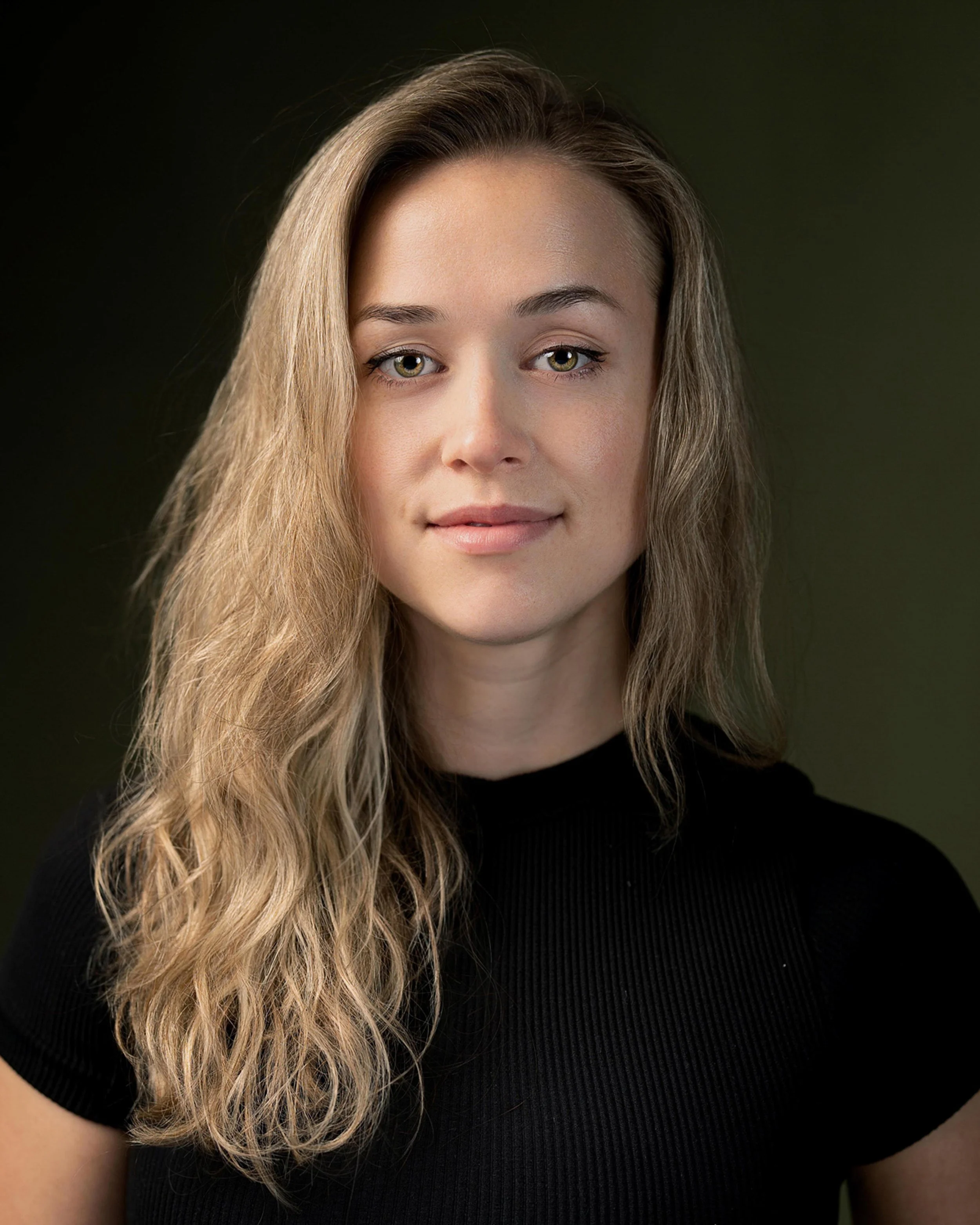Close-up of a young woman with long, wavy blonde hair wearing a black top, looking into the camera with a neutral expression against a dark background.