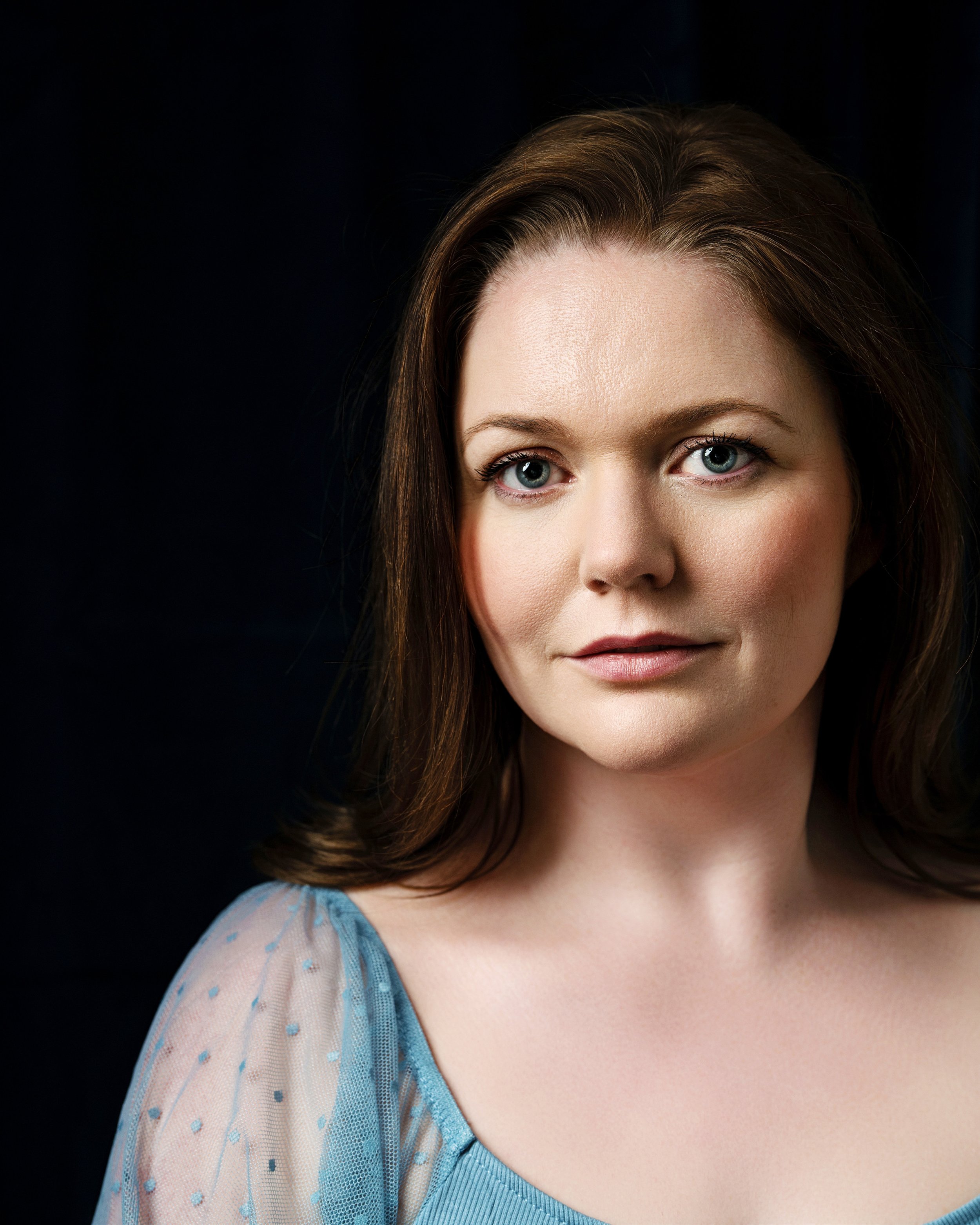 Headshot of a woman in a blue blouse with blue eyes in front of a black backdrop