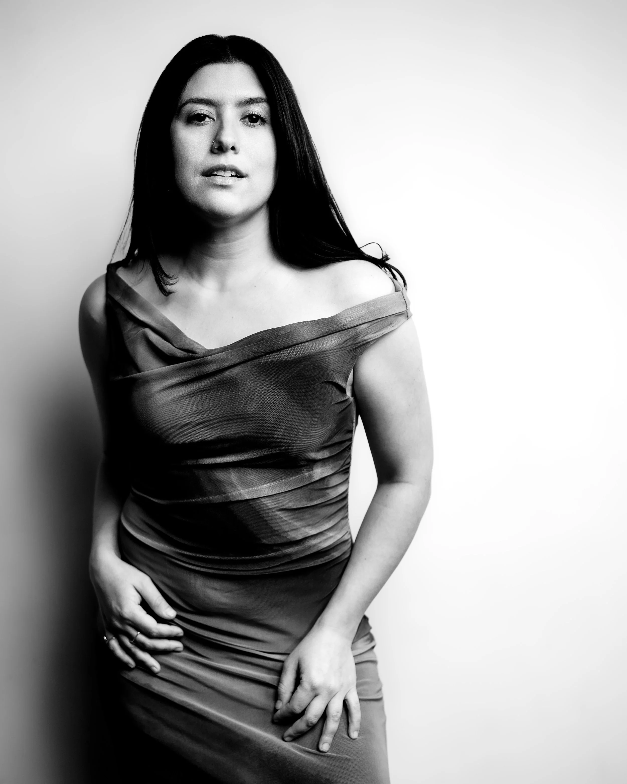 Black and white photo of a woman with long dark hair, wearing a dress, posing with one hand on her thigh and looking at the camera.