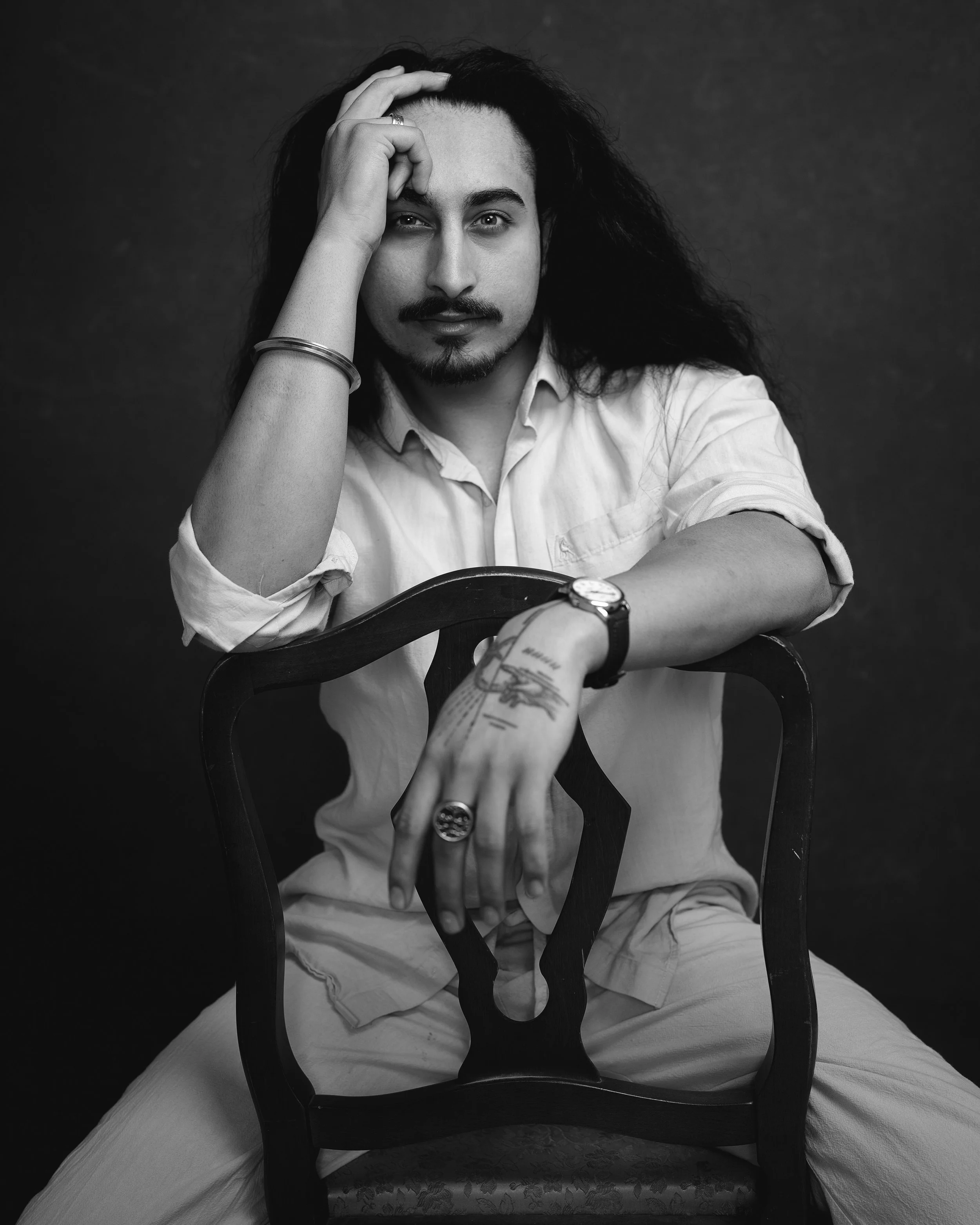 Black and white portrait of a man with long hair, beard, and mustache, sitting on a chair with a dark background, wearing a white shirt, with one hand on his forehead and the other on the back of the chair, looking directly at the camera.