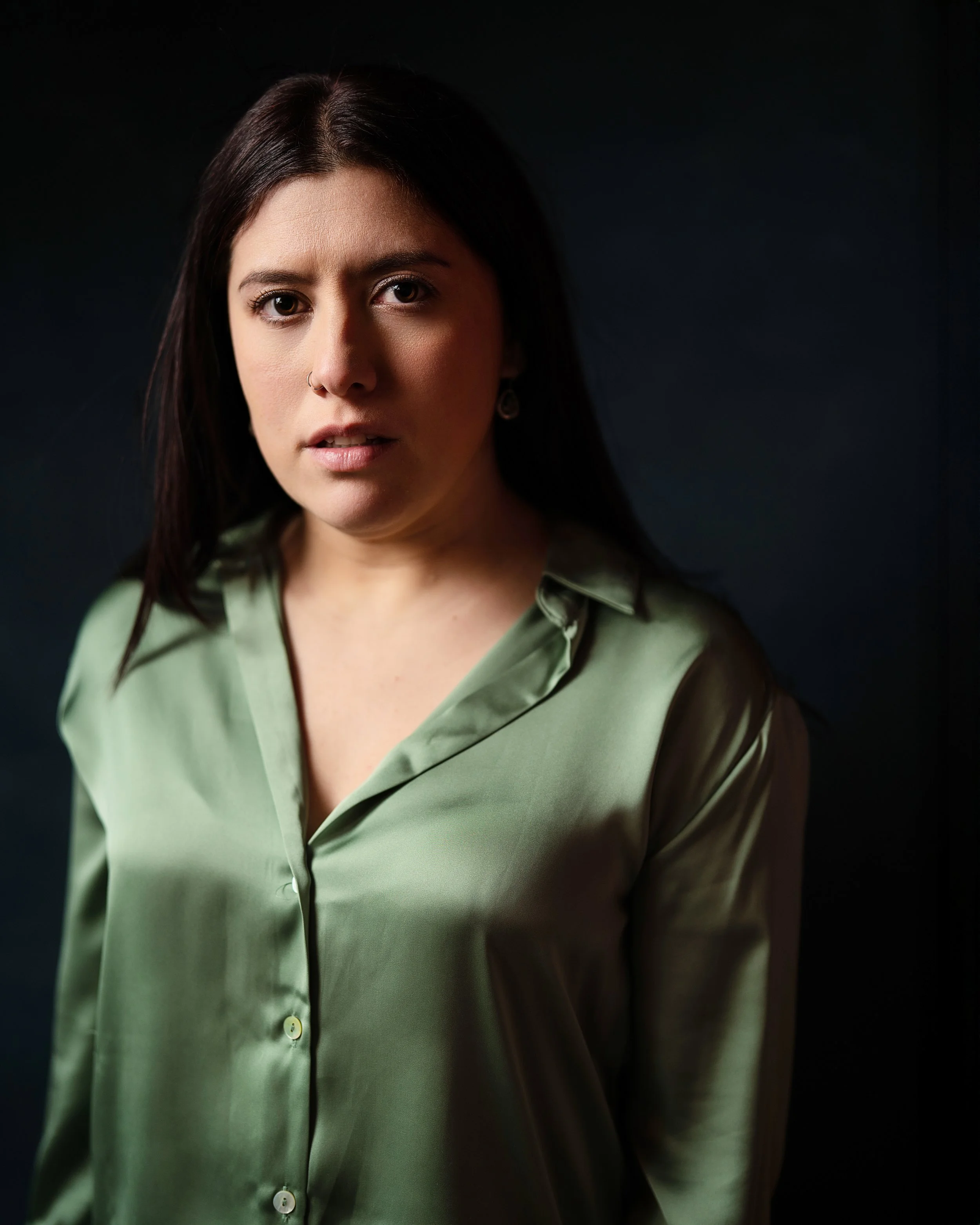 A woman with dark brown hair, wearing a green button-up shirt, and a nose ring, looking directly at the camera against a dark background.