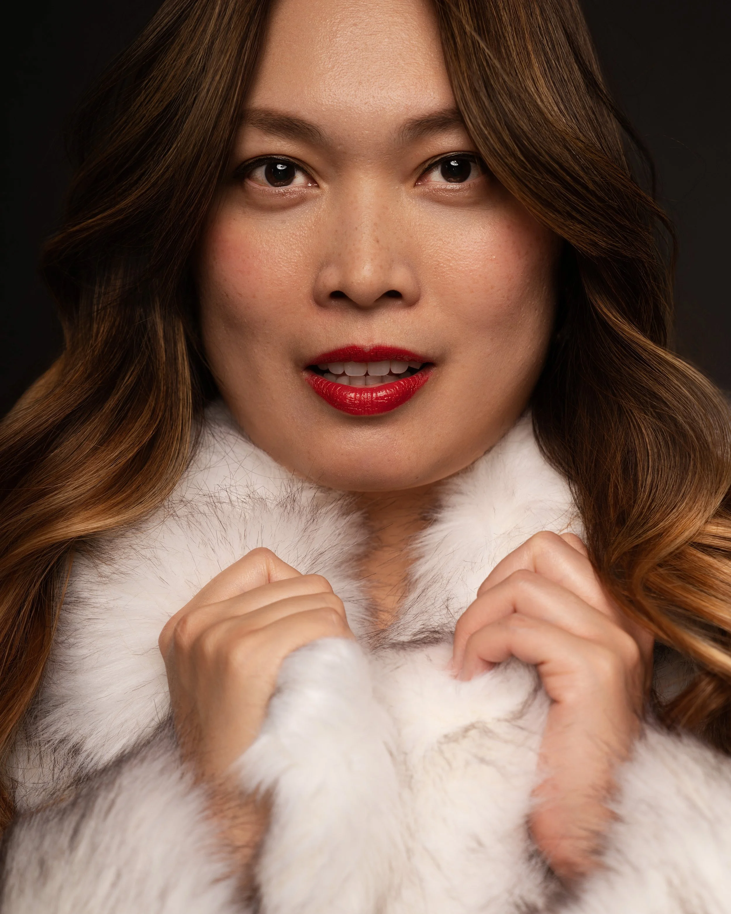 Close-up portrait of a woman with wavy brown hair and red lipstick, holding a piece of white faux fur fabric.