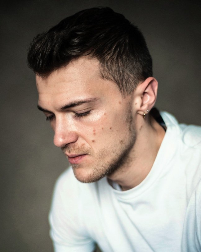 Headshot of a man with eyes closed in a white t-shirt in front of am olive green backdrop