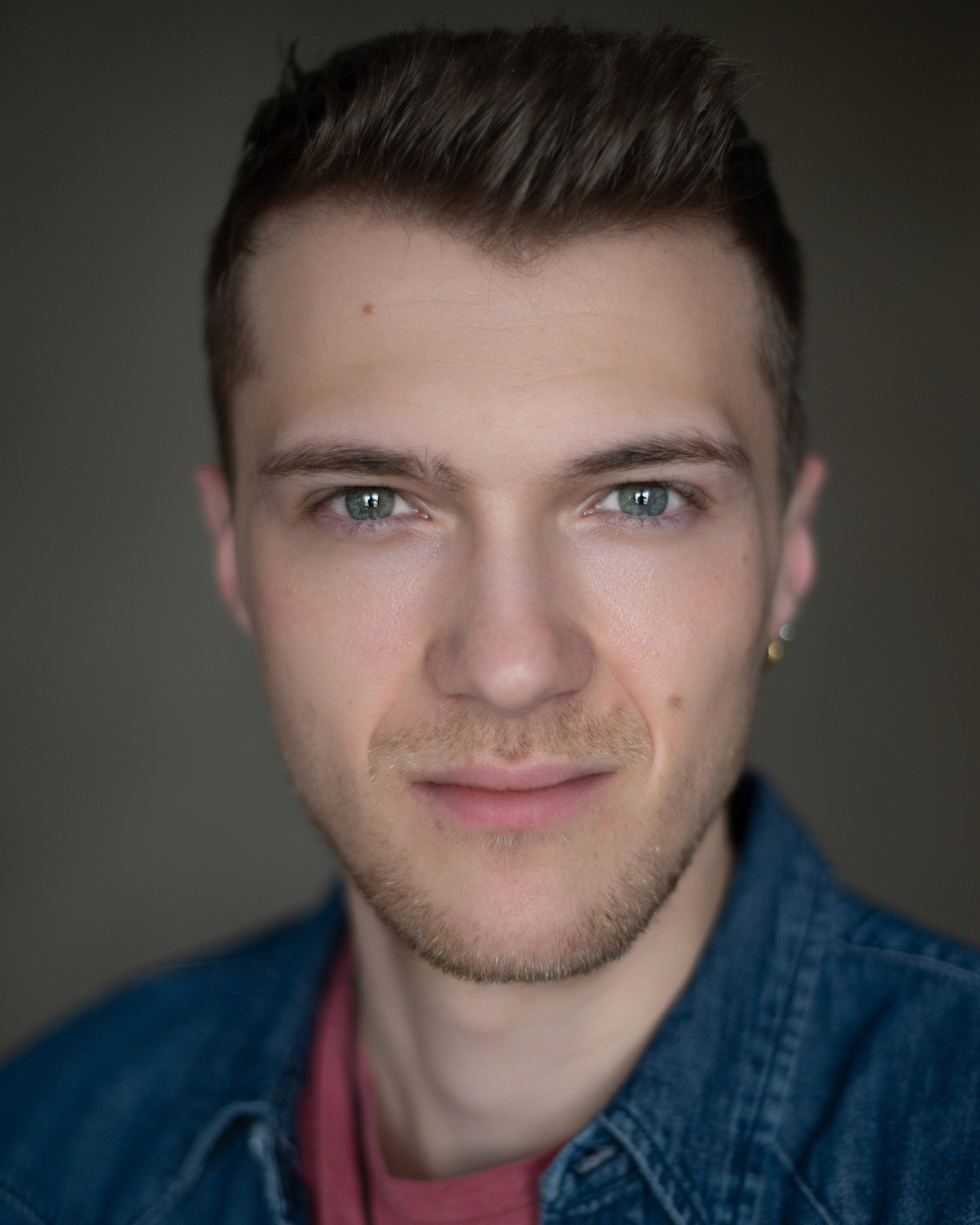 Headshot of a man in a red t-shirt and blue jean button up with crystal blue green eyes in front of an olive green backdrop