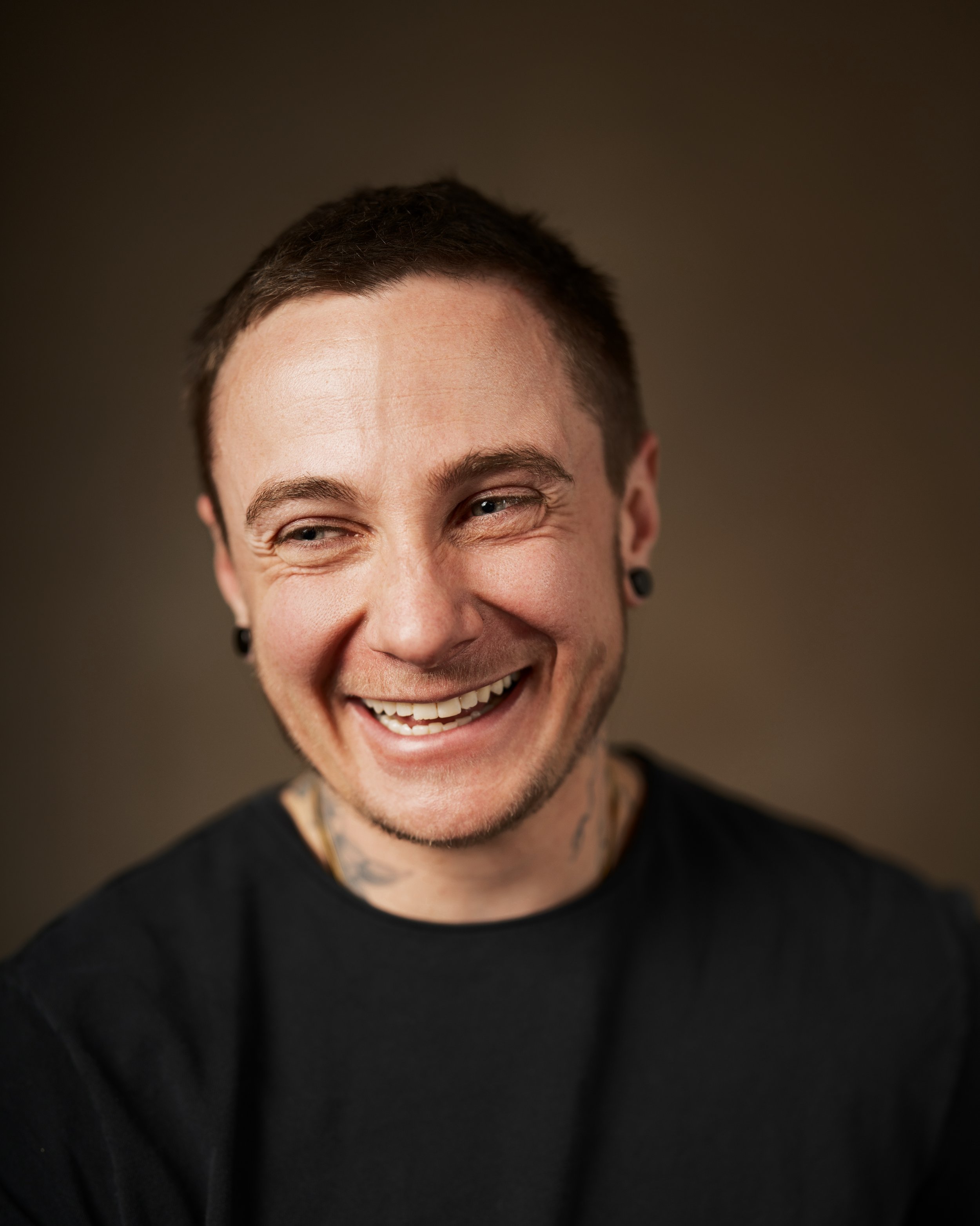 Headshot of a man laughing wearing a black t-shirt with a kaki backdrop