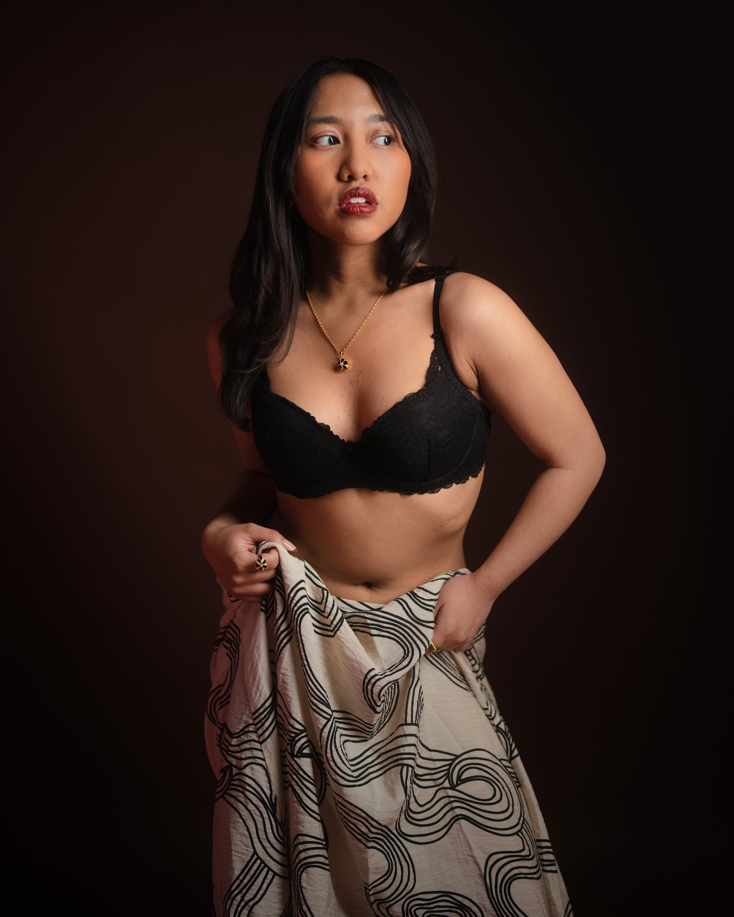 A young woman with long dark hair wearing a black lace bra, a gold necklace with a clover pendant, and a flowing skirt with swirling black lines on a white background, posing against a dark background.