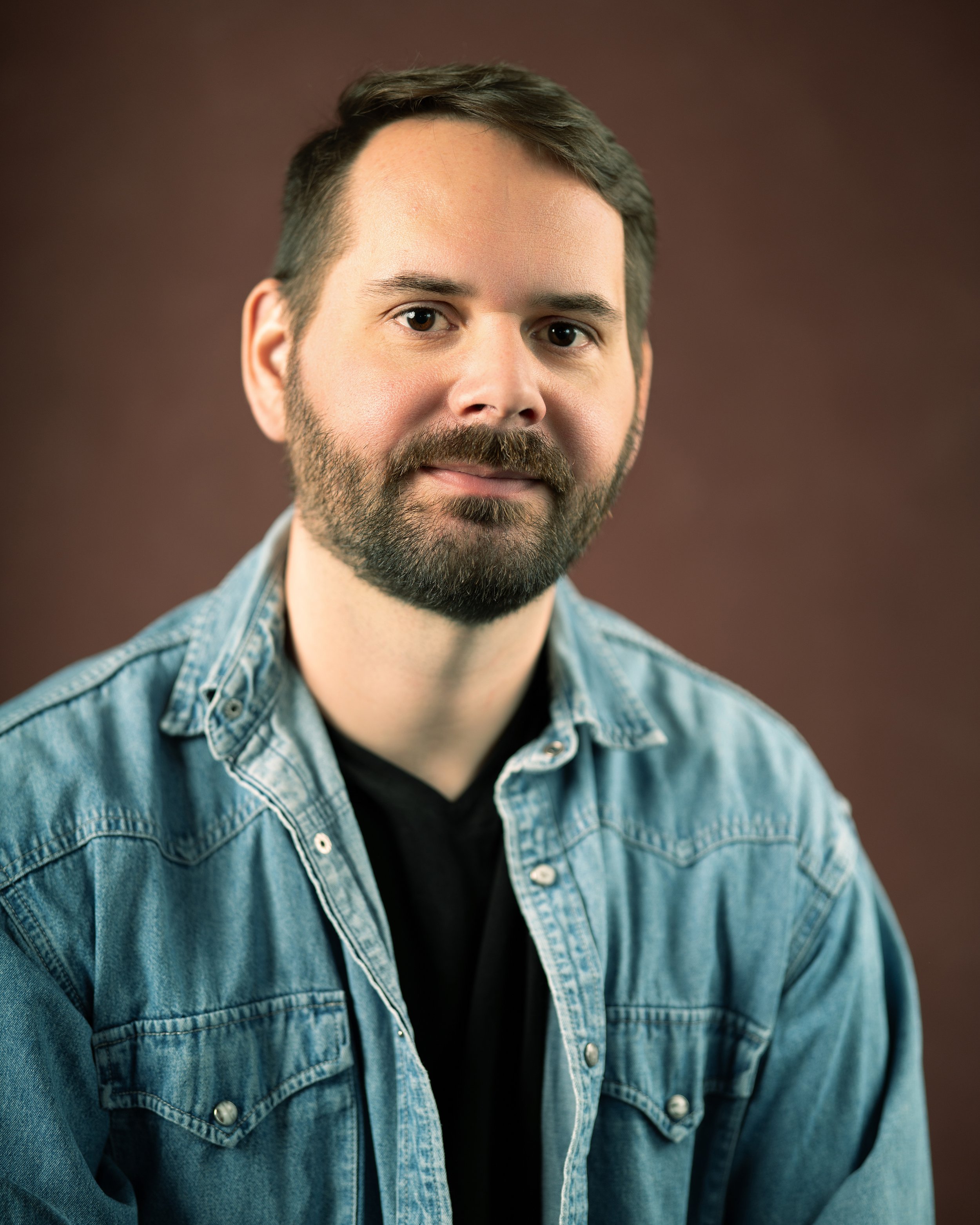 Headshot of a man in a black t-shirt and blu jean button up shirt in front of a red backdrop
