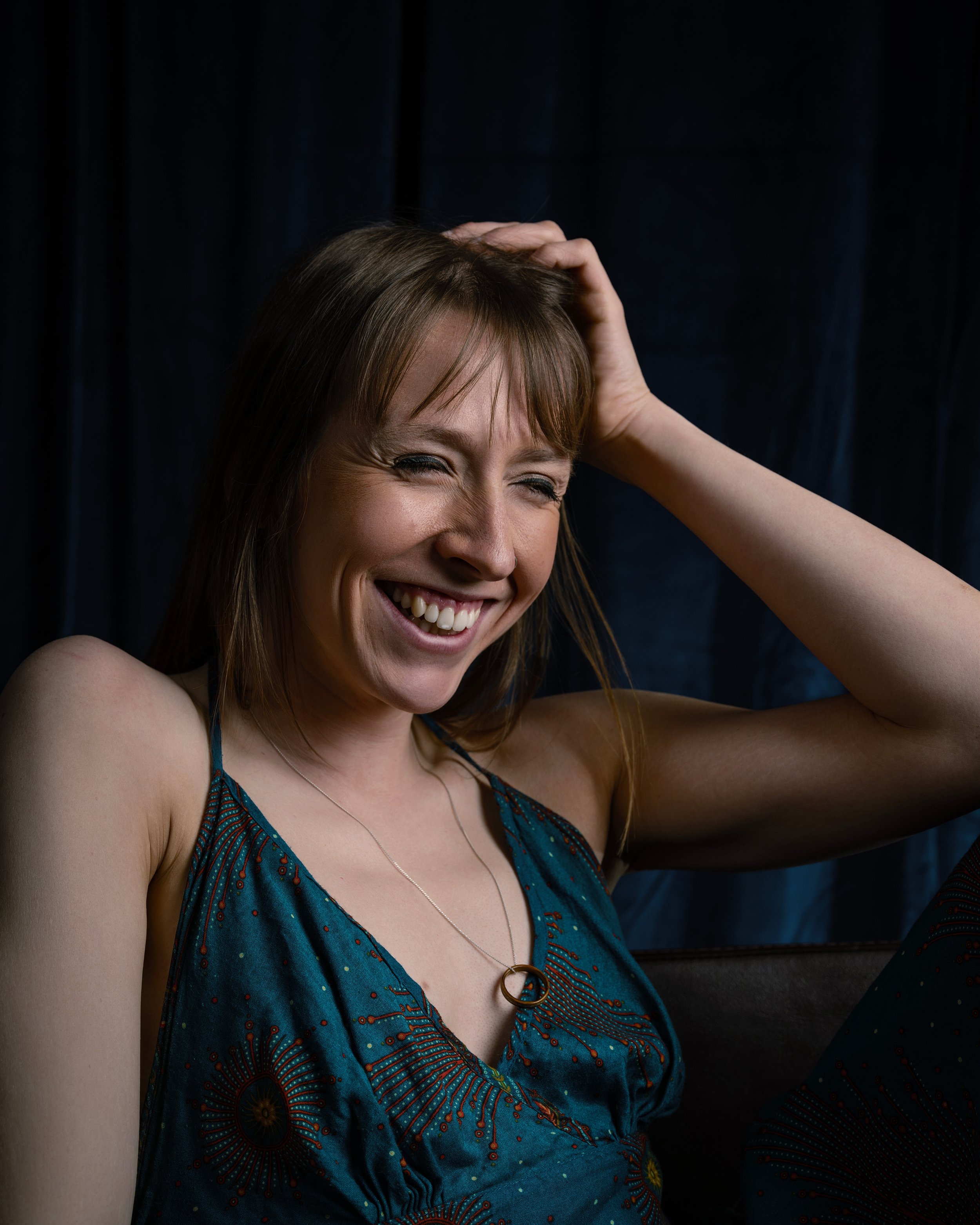 Headshot of a woman laughing in a turquoise dress holding her head in front of a blue backdrop