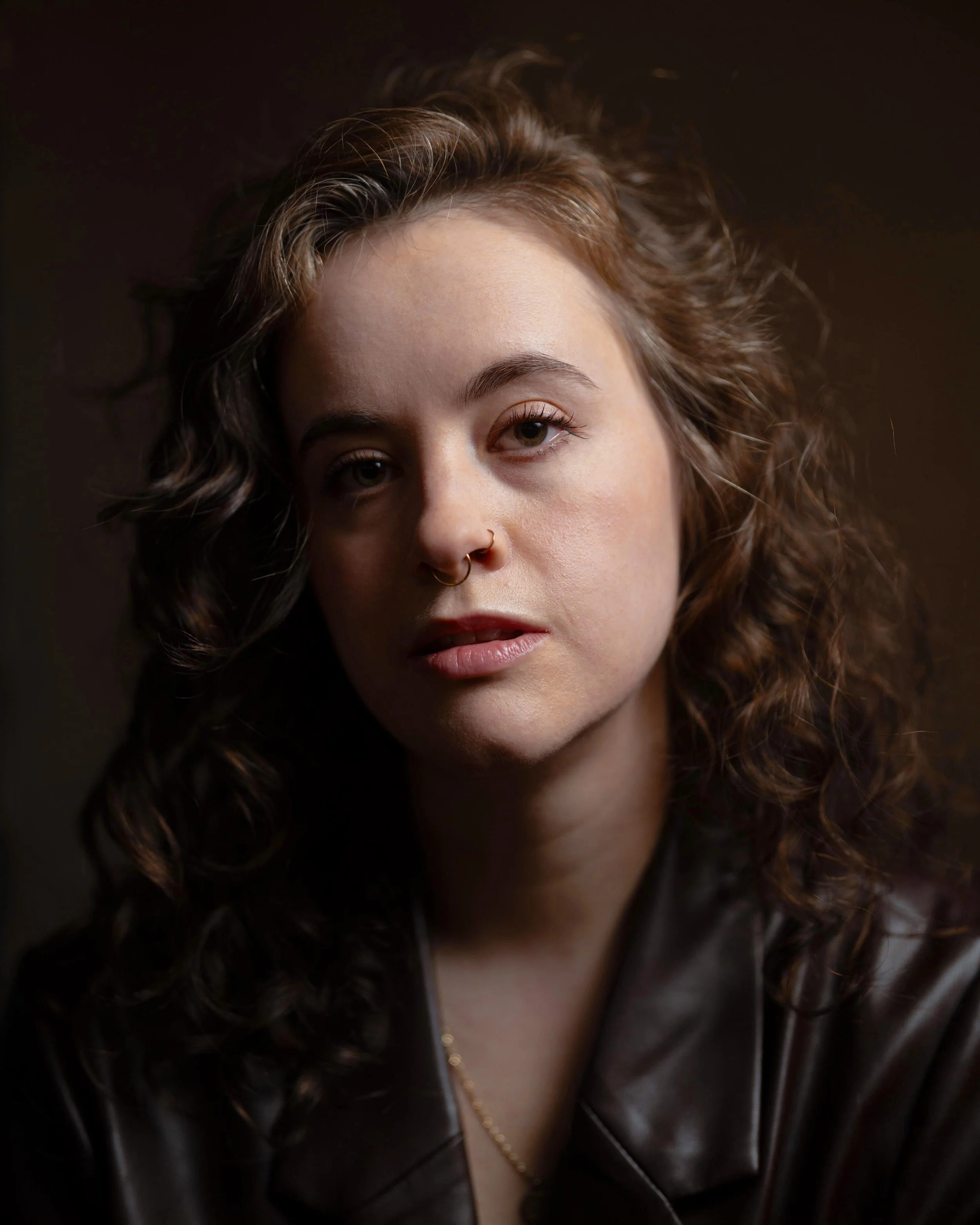 A young woman with curly brown hair, gold hoop nose rings, wearing a dark leather jacket, and a necklace. The image is a close-up portrait with a dark background.