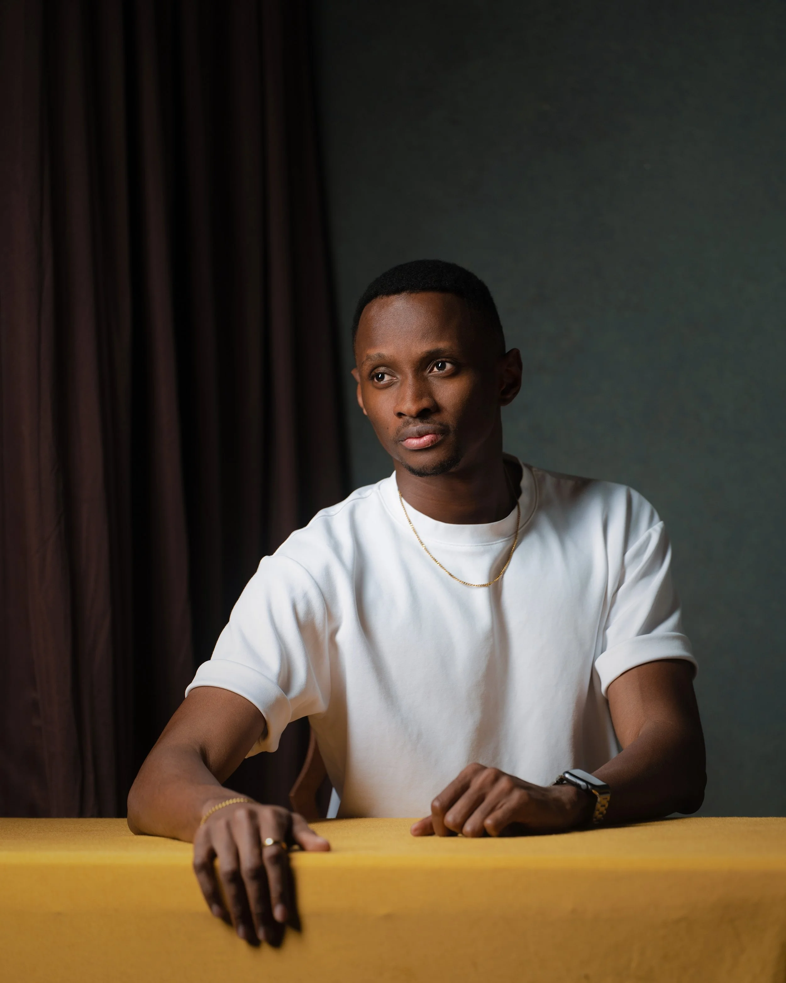 A young man with short hair, wearing a white t-shirt, a gold chain, and a smartwatch, sitting at a yellow table, with dark curtains and a gray wall in the background.