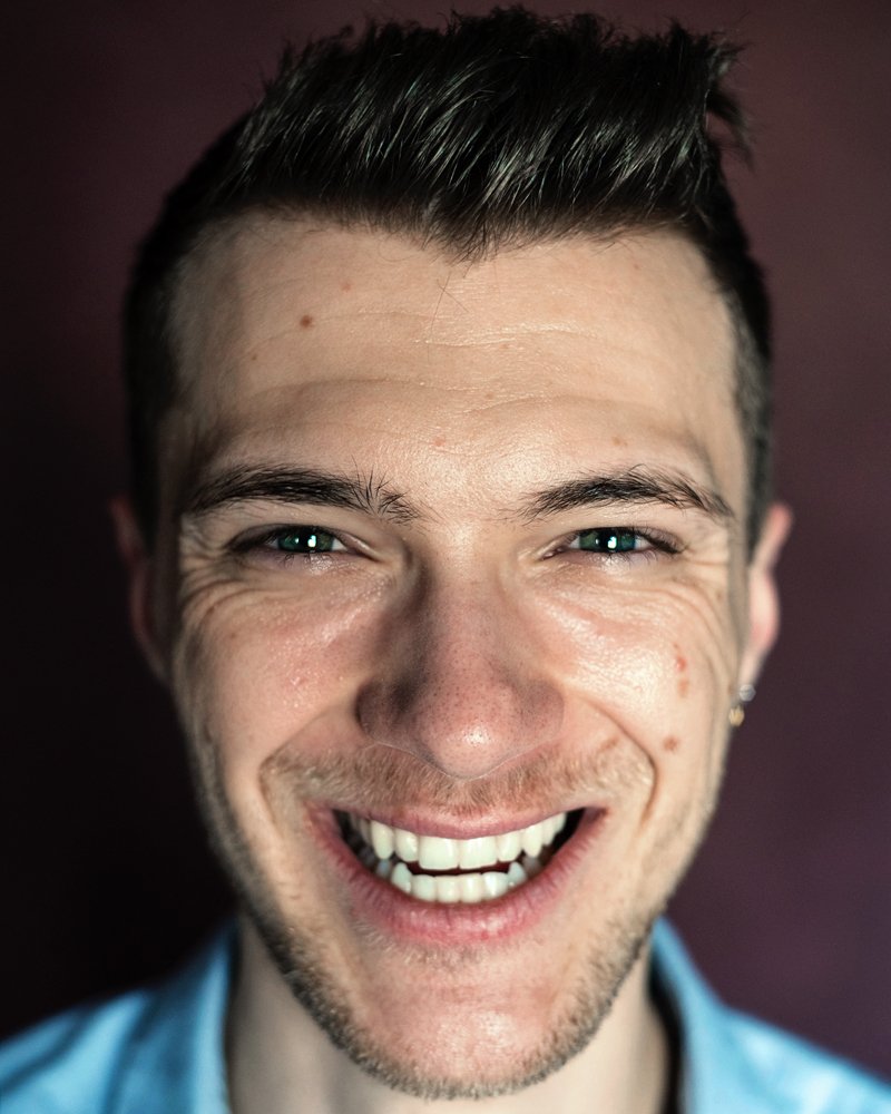 Headshot of a man with a big smile in a blue shirt in front of a red backdrop