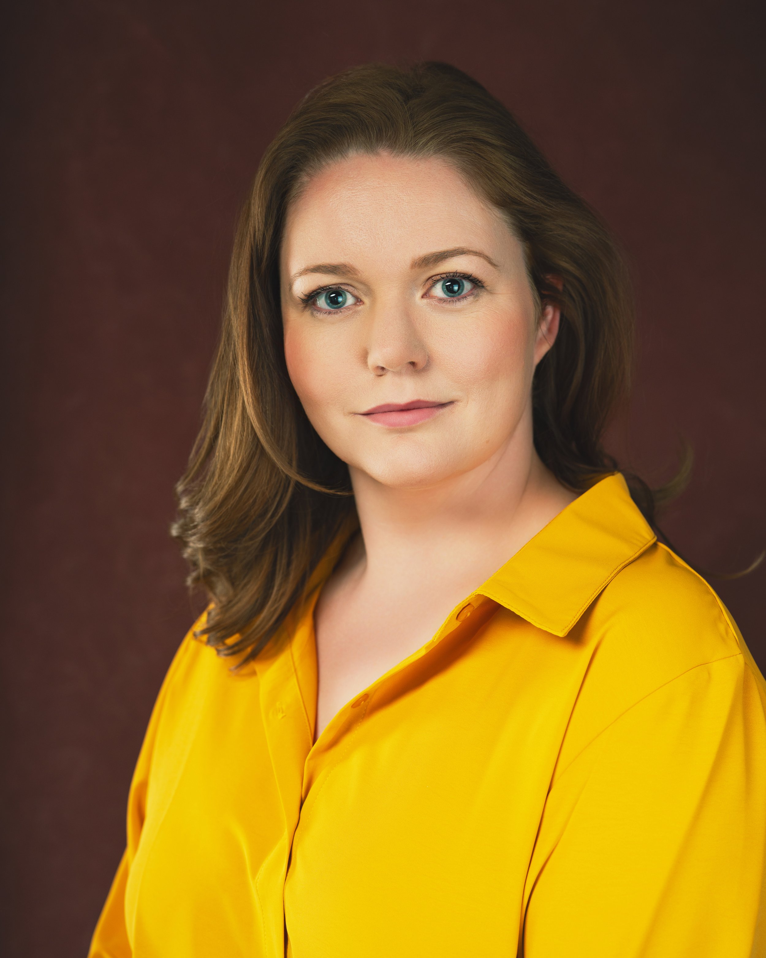 Headshot of a woman with brown hair in a yellow shirt in front of a red backdrop