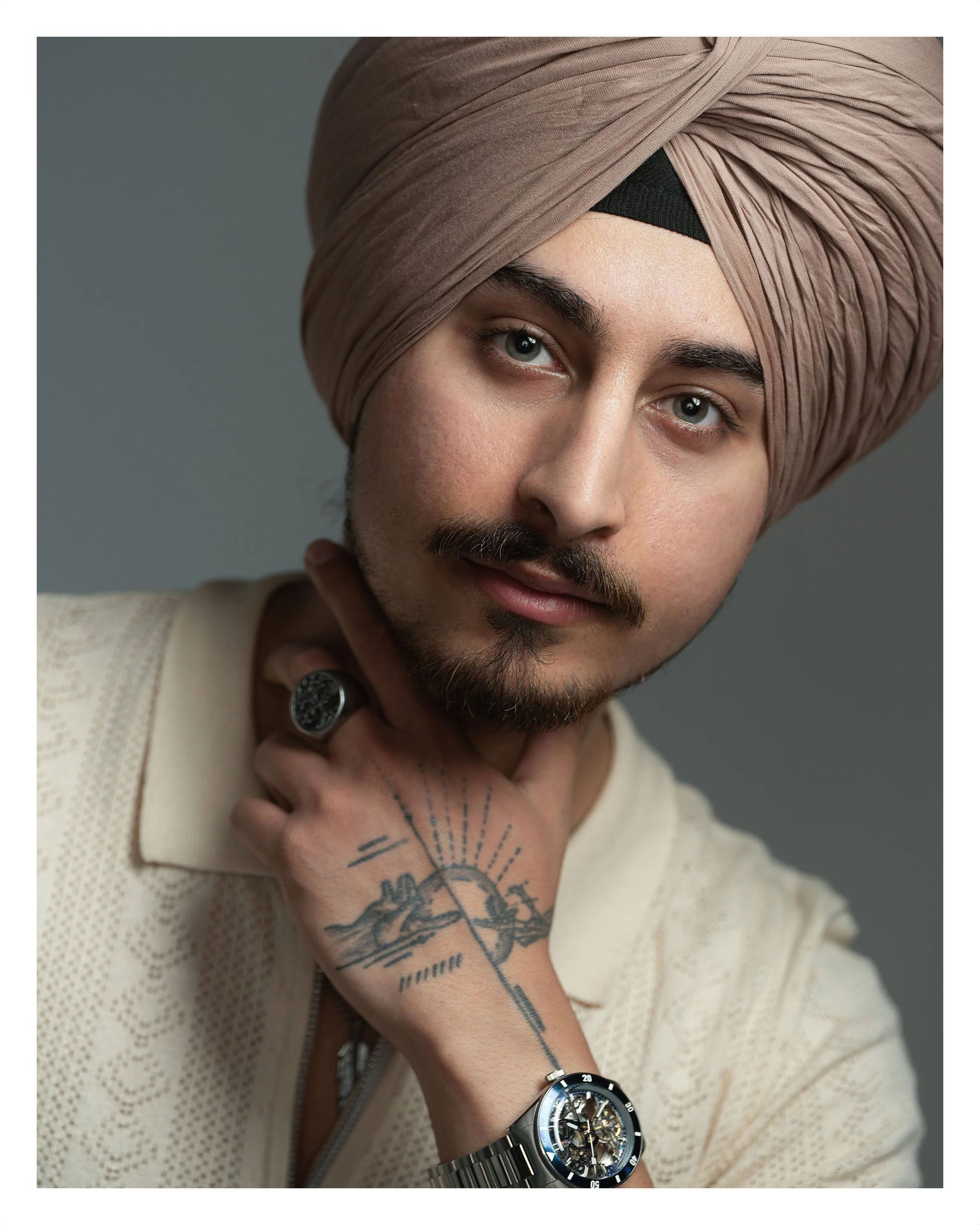 A man wearing a beige turban and a beige sweater, with a beard and blue eyes, is resting his chin on his hand. He wears a large black watch and a silver bracelet.