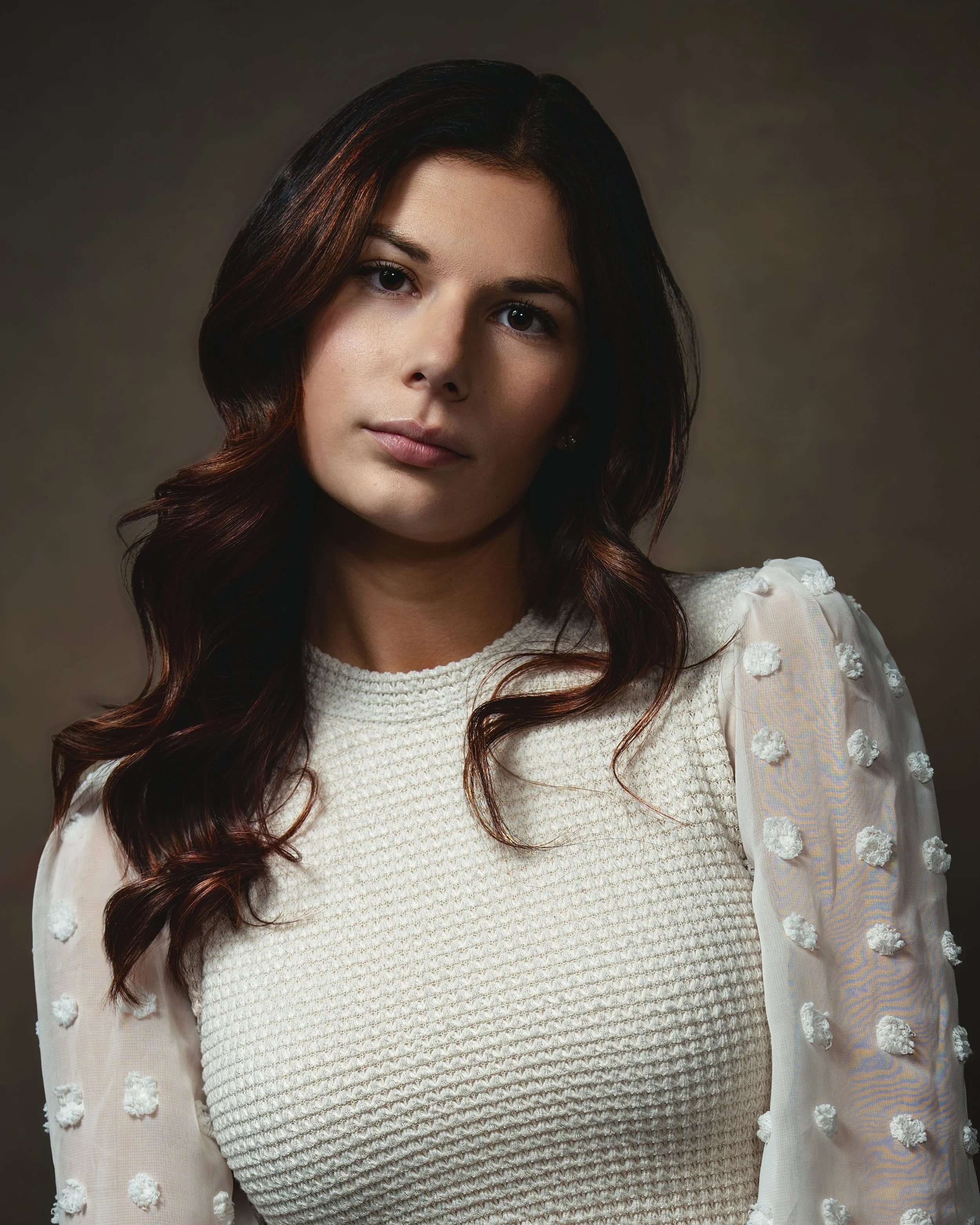 A young woman with wavy dark brown hair and fair skin looks at the camera with a neutral expression. She is wearing a white textured dress with a knitted bodice and a sheer white sleeve with 3D floral embroidery.