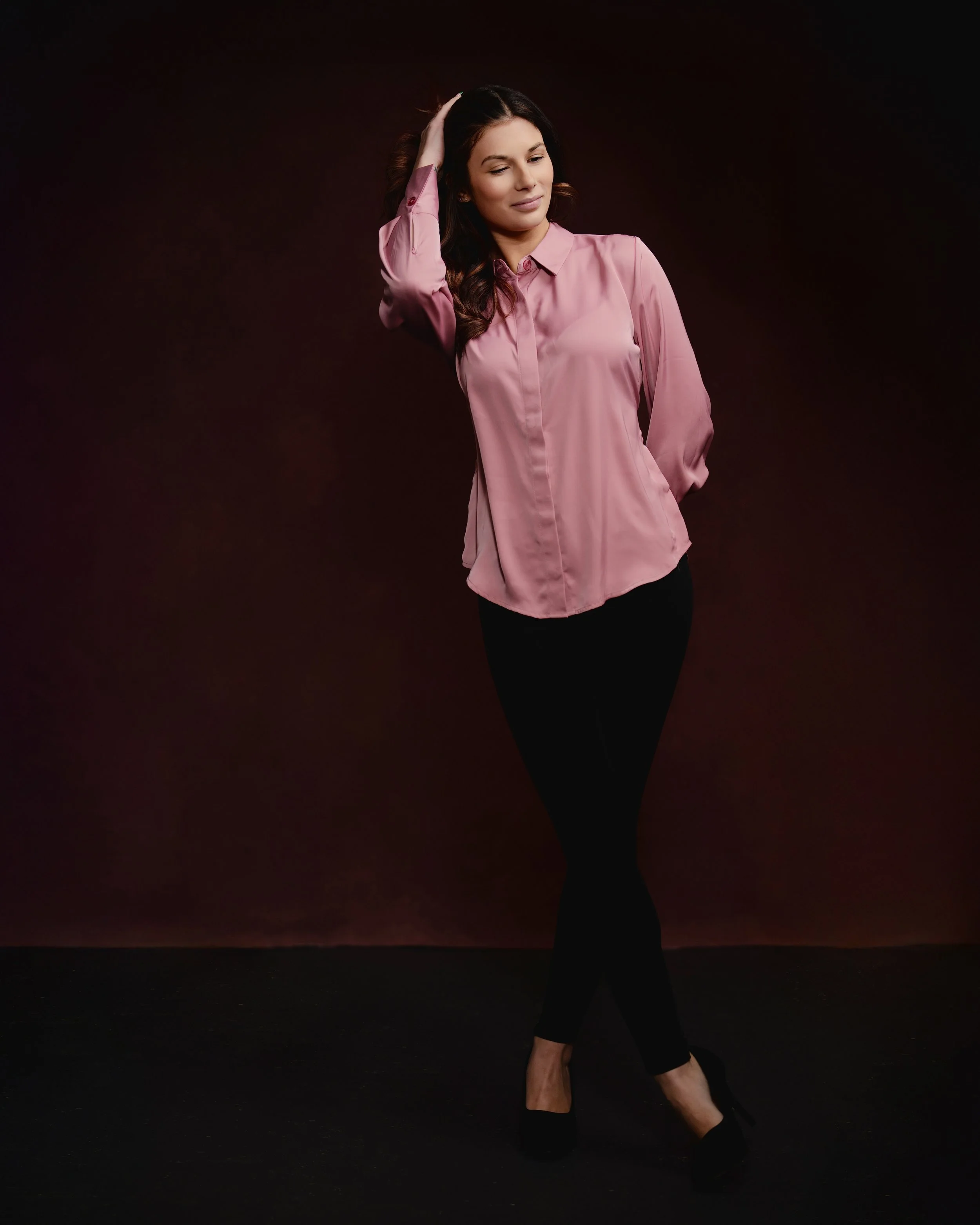 A woman in a pink satin shirt and black pants stands against a dark background, with her eyes closed and one hand on her head.