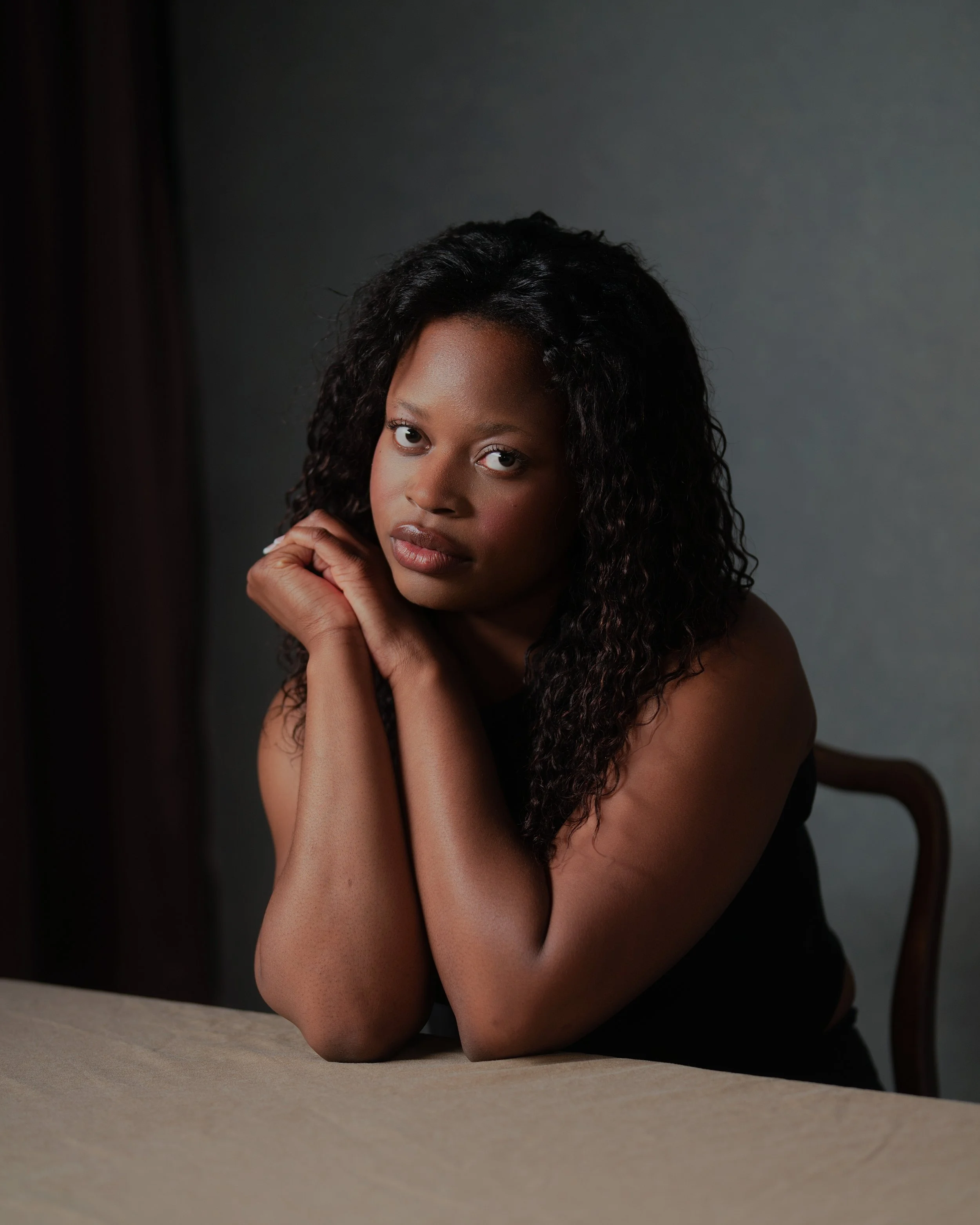 A woman with curly black hair and brown skin sitting at a table, resting her chin on her hand, looking directly at the camera with a serious expression against a gray background.