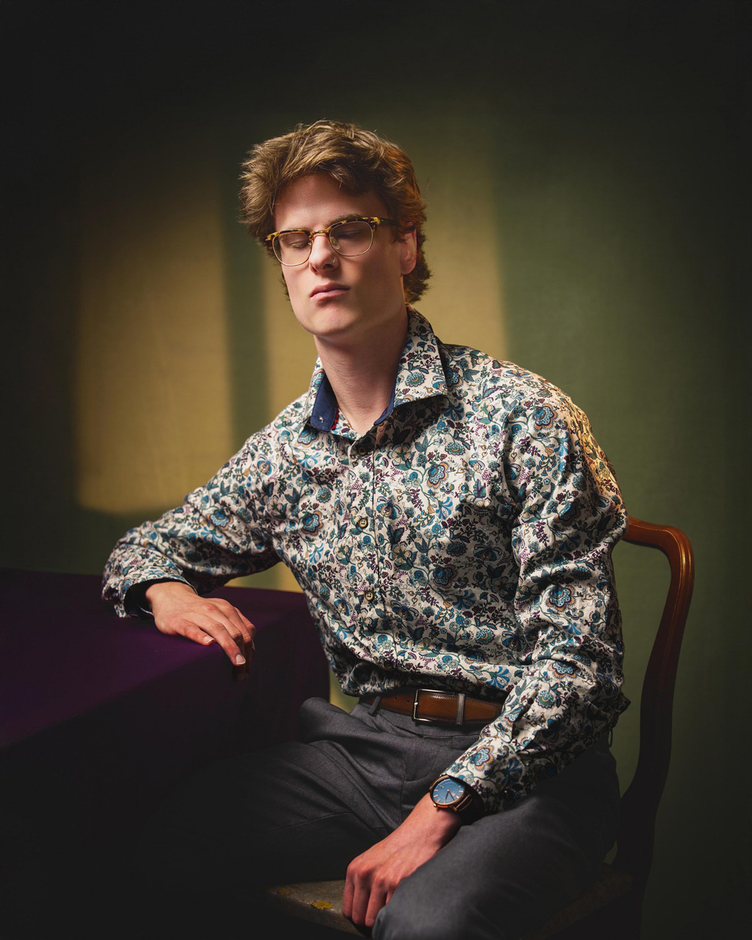 A young man with wavy brown hair and glasses, wearing a floral patterned shirt, seated with a calm expression, resting his arm on a dark table, and wearing a watch on his wrist.