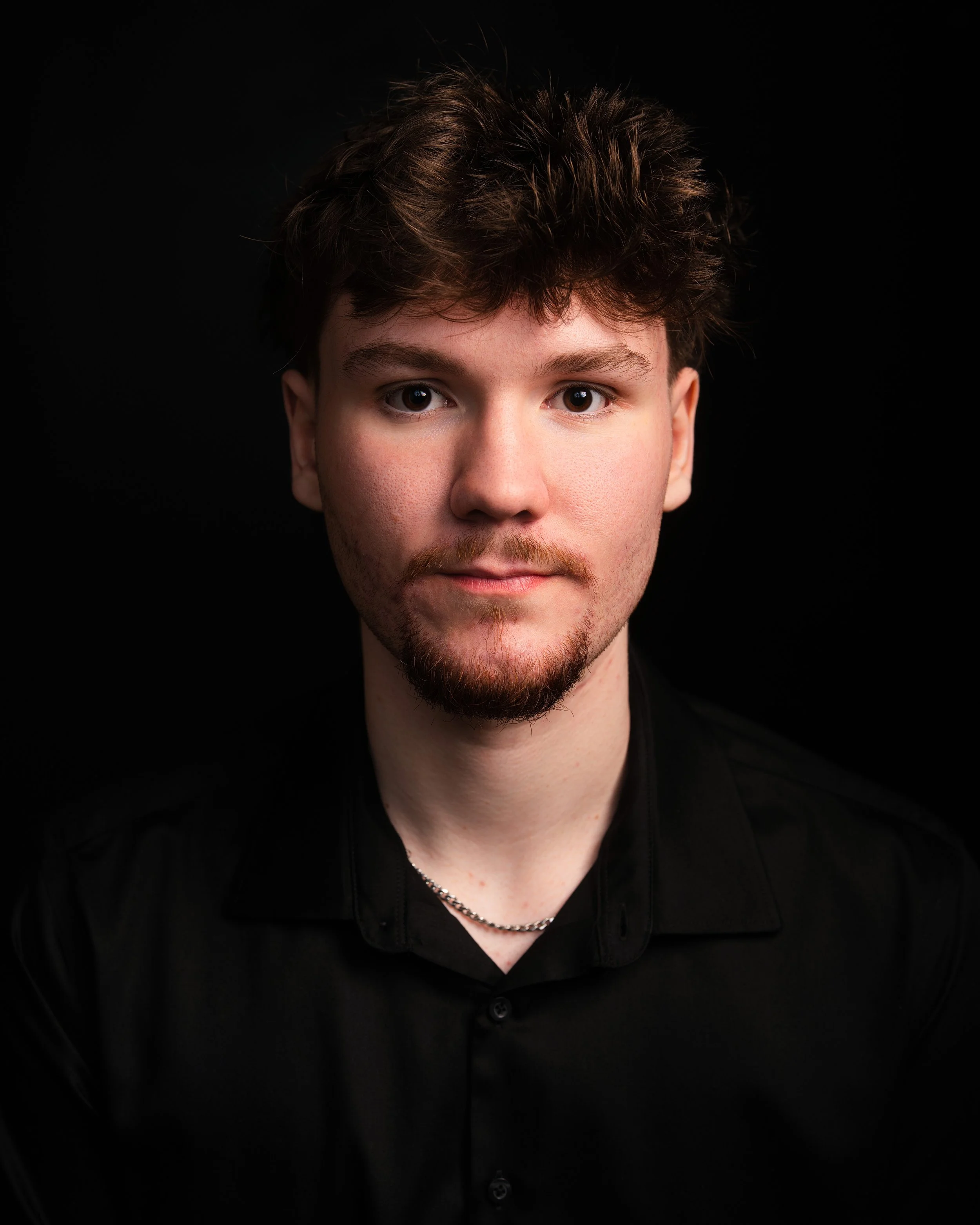 Close-up portrait of a young man with brown curly hair, facial hair, and light skin, wearing a black shirt and silver chain necklace, against a black background.