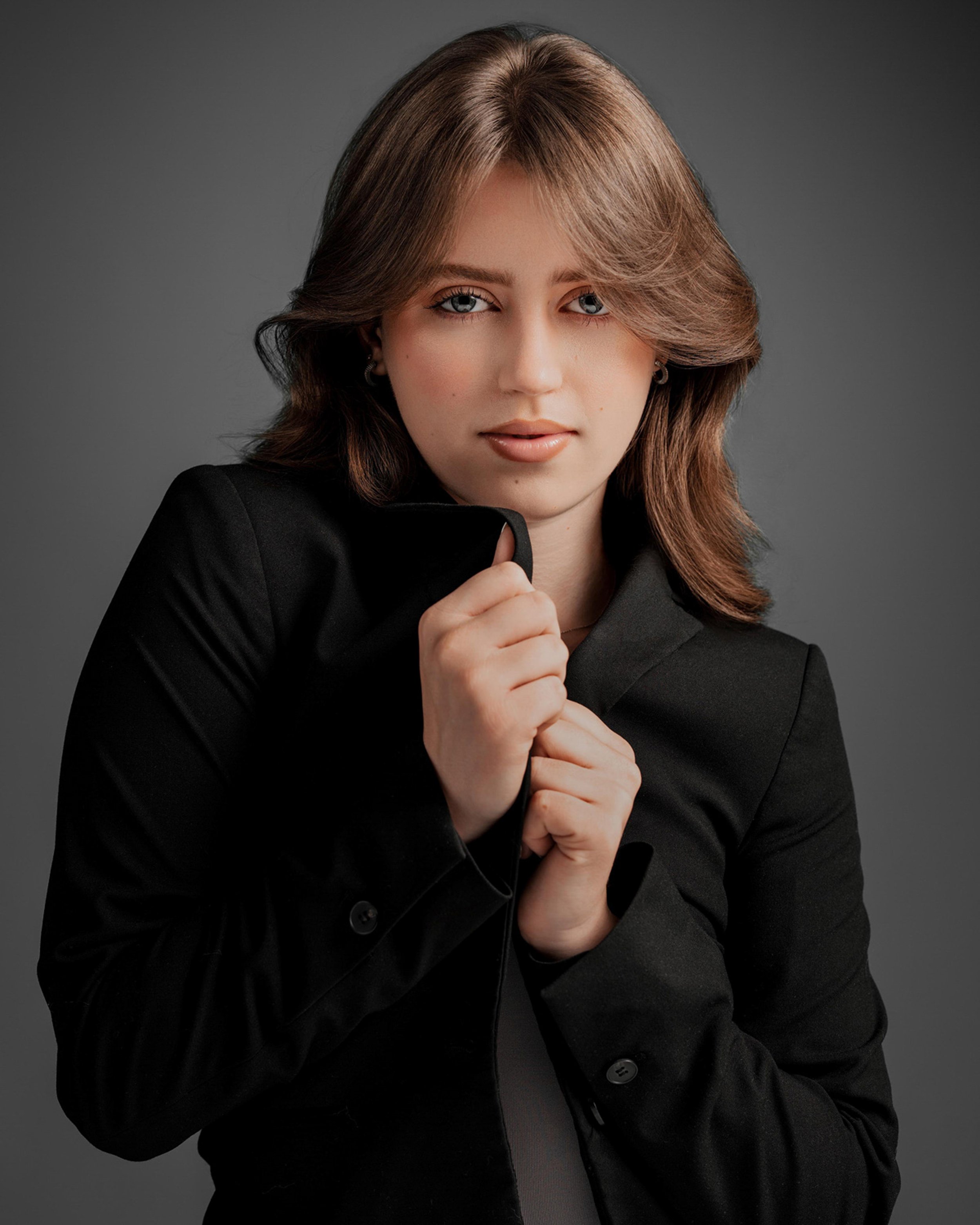 Portrait of a young woman with shoulder-length brown hair, blue eyes, and light makeup, wearing a black blazer, against a dark gray background.
