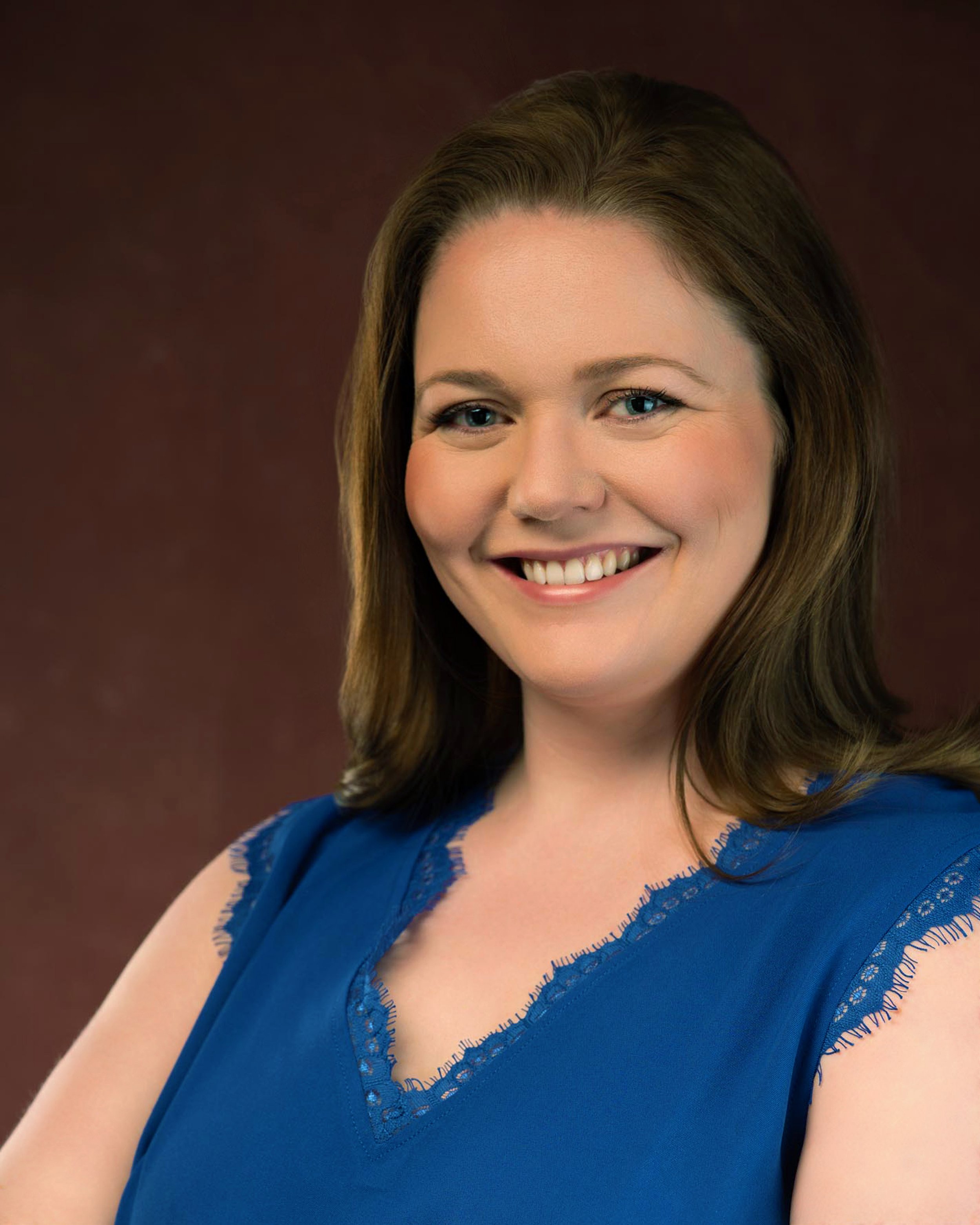 Headshot of a woman in a blue blouse with brown hair and blue eyes in front of a red backdrop