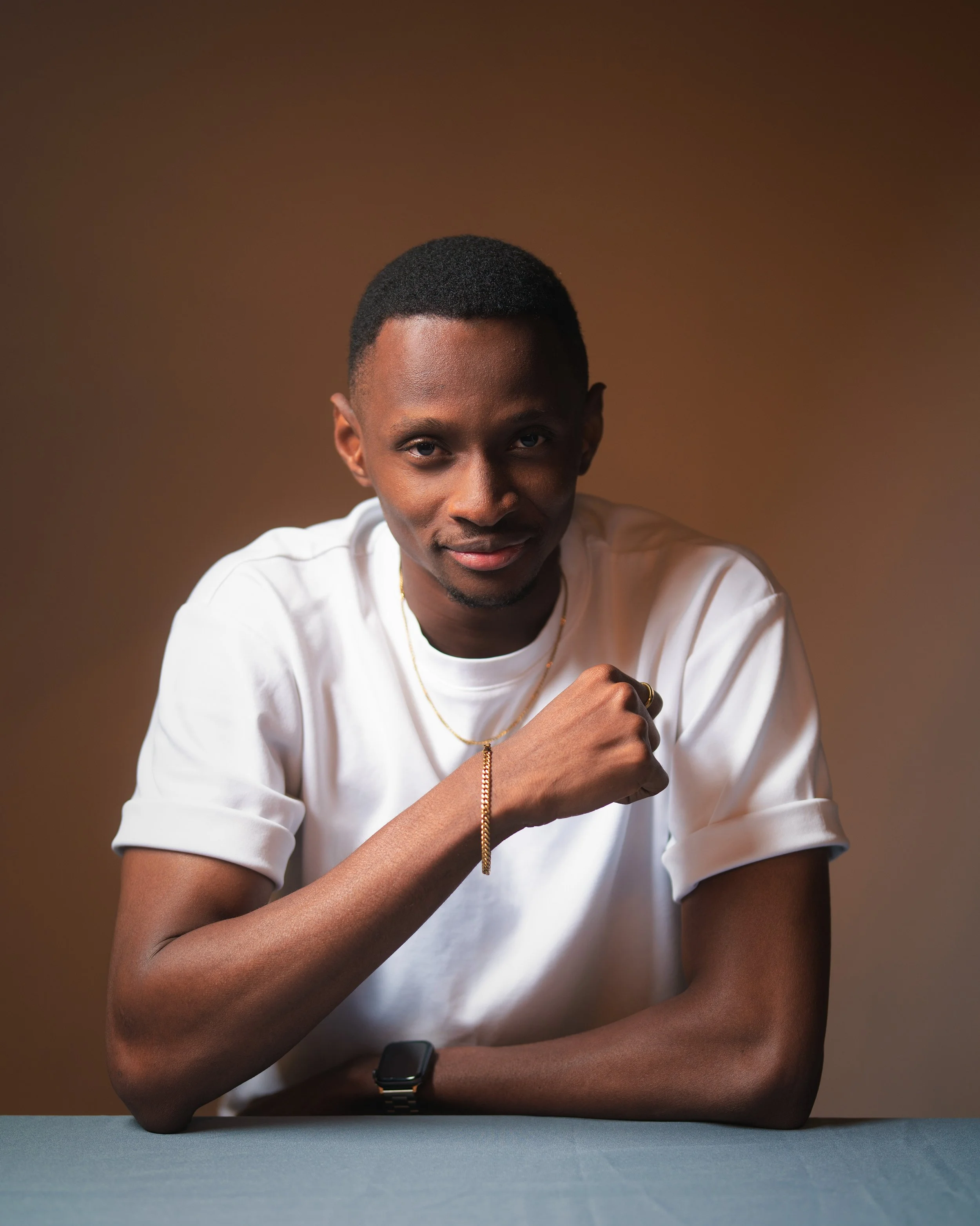 Close-up of a young man with short hair wearing a white t-shirt, gold jewelry, and a smartwatch, seated at a table, with a warm brown background.