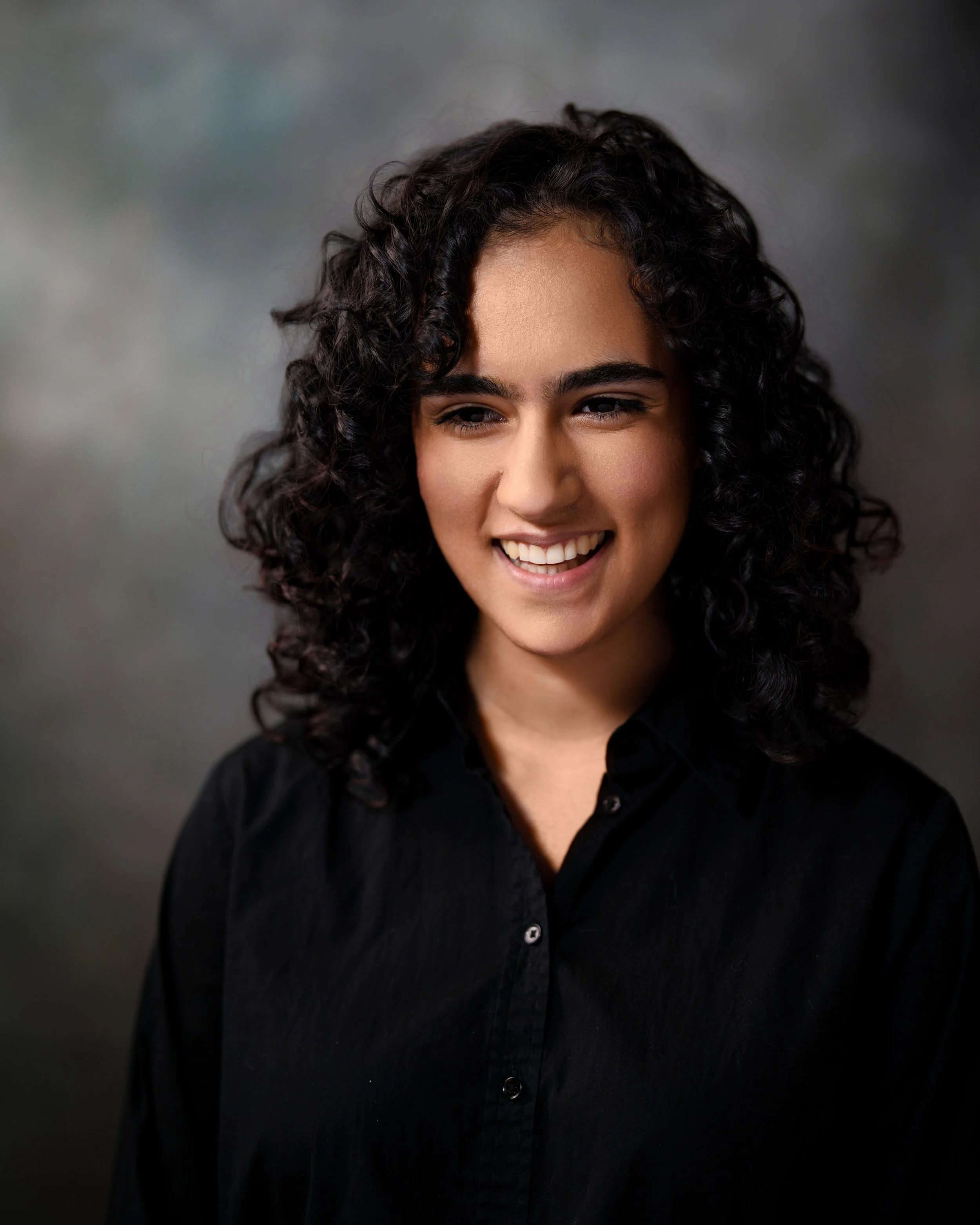 Close-up portrait of a woman with curly dark hair and a smile, wearing a black shirt, against a blurred gray background.