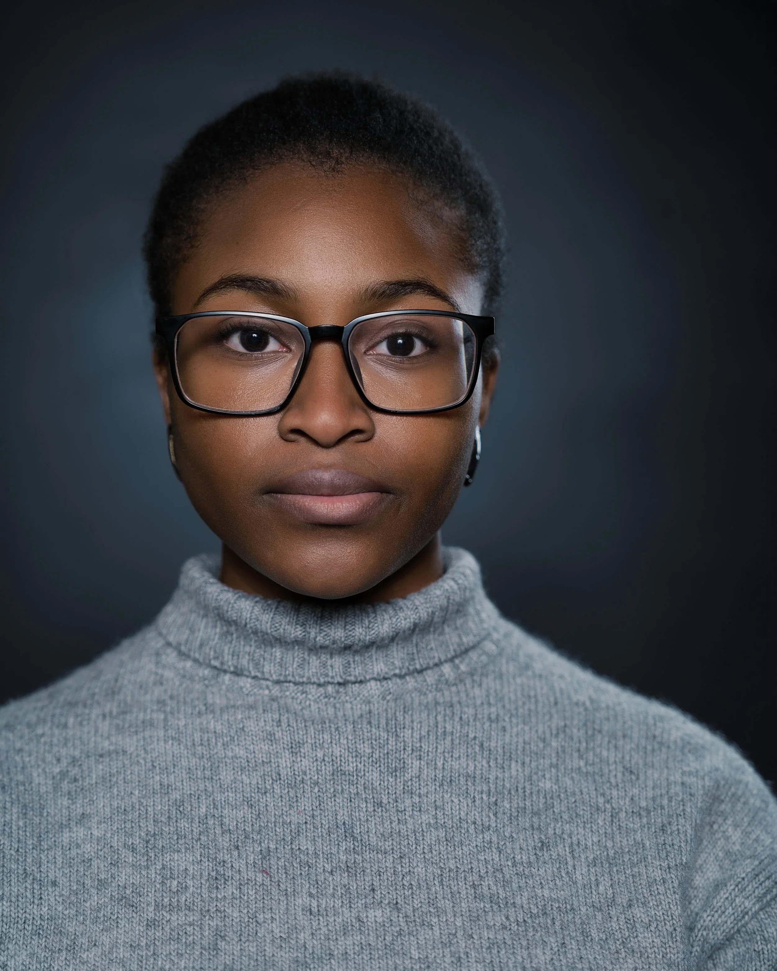 Close-up of a woman with glasses wearing a gray turtleneck sweater and dark background.