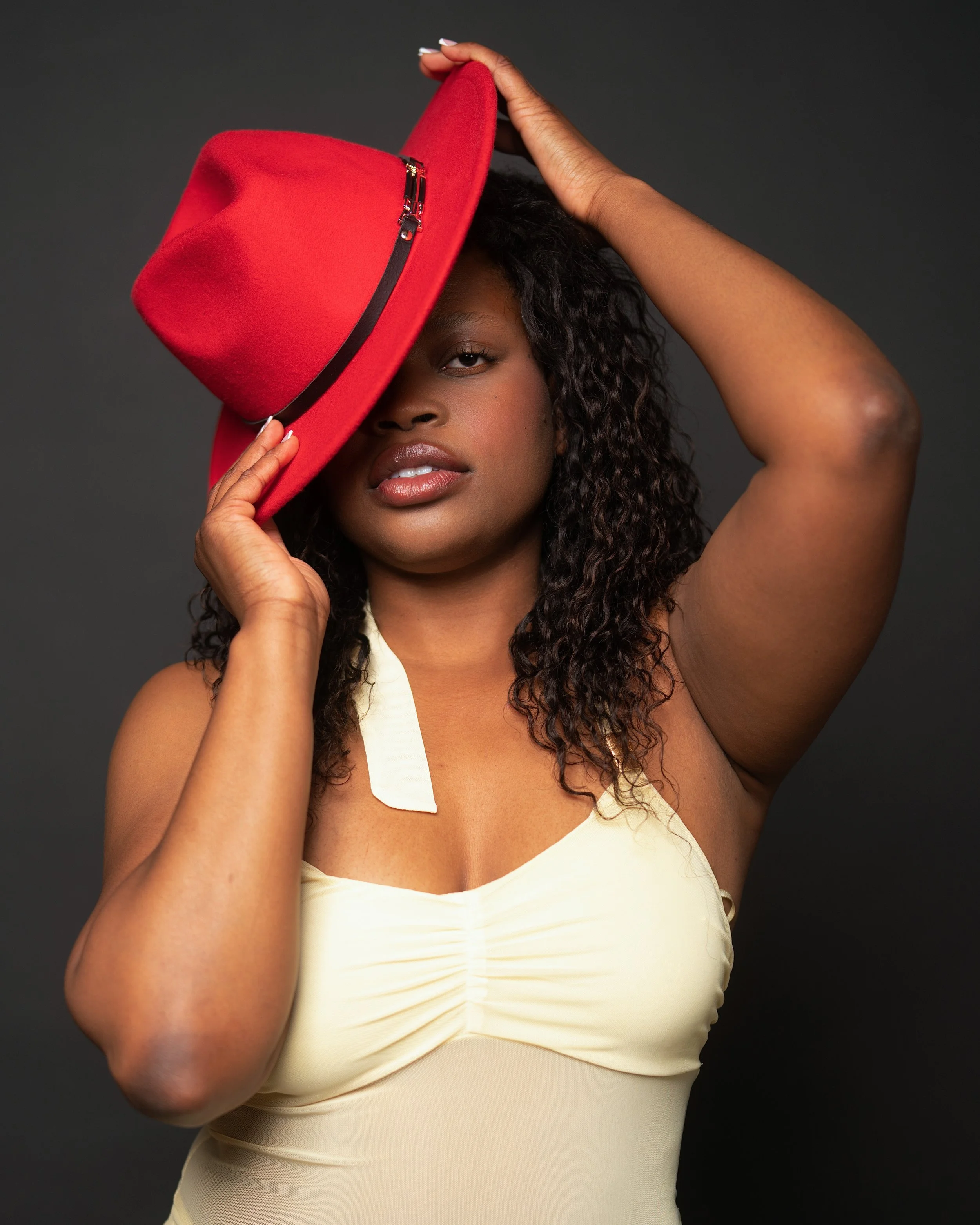 A woman with dark curly hair, wearing a cream-colored dress, holds a red hat with a black band over her eyes, against a dark background.