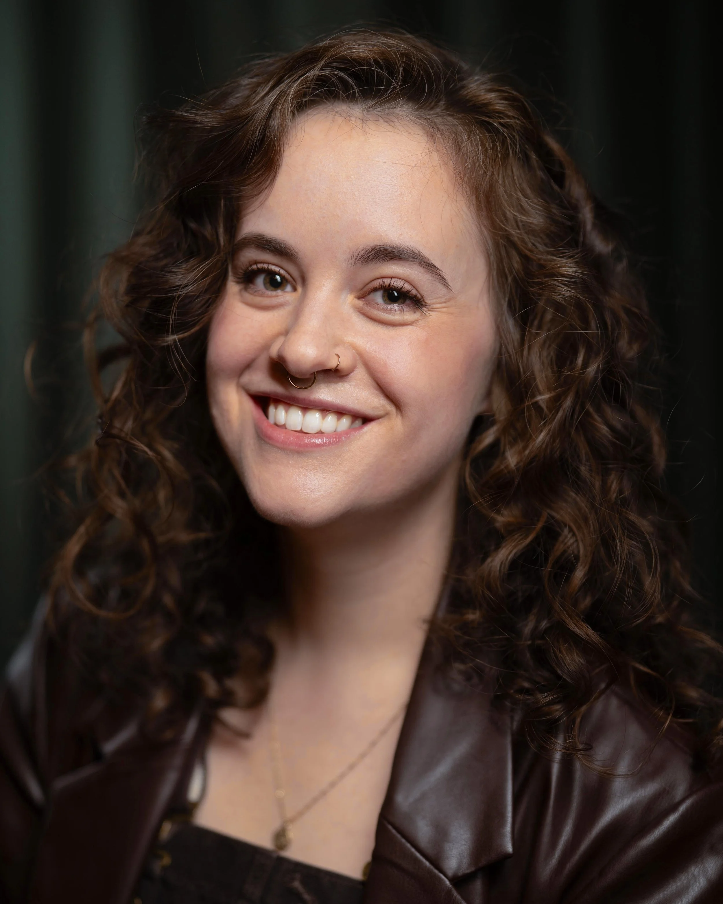 Close-up of a woman with curly brown hair smiling, wearing a leather jacket and a gold necklace with a small pendant.