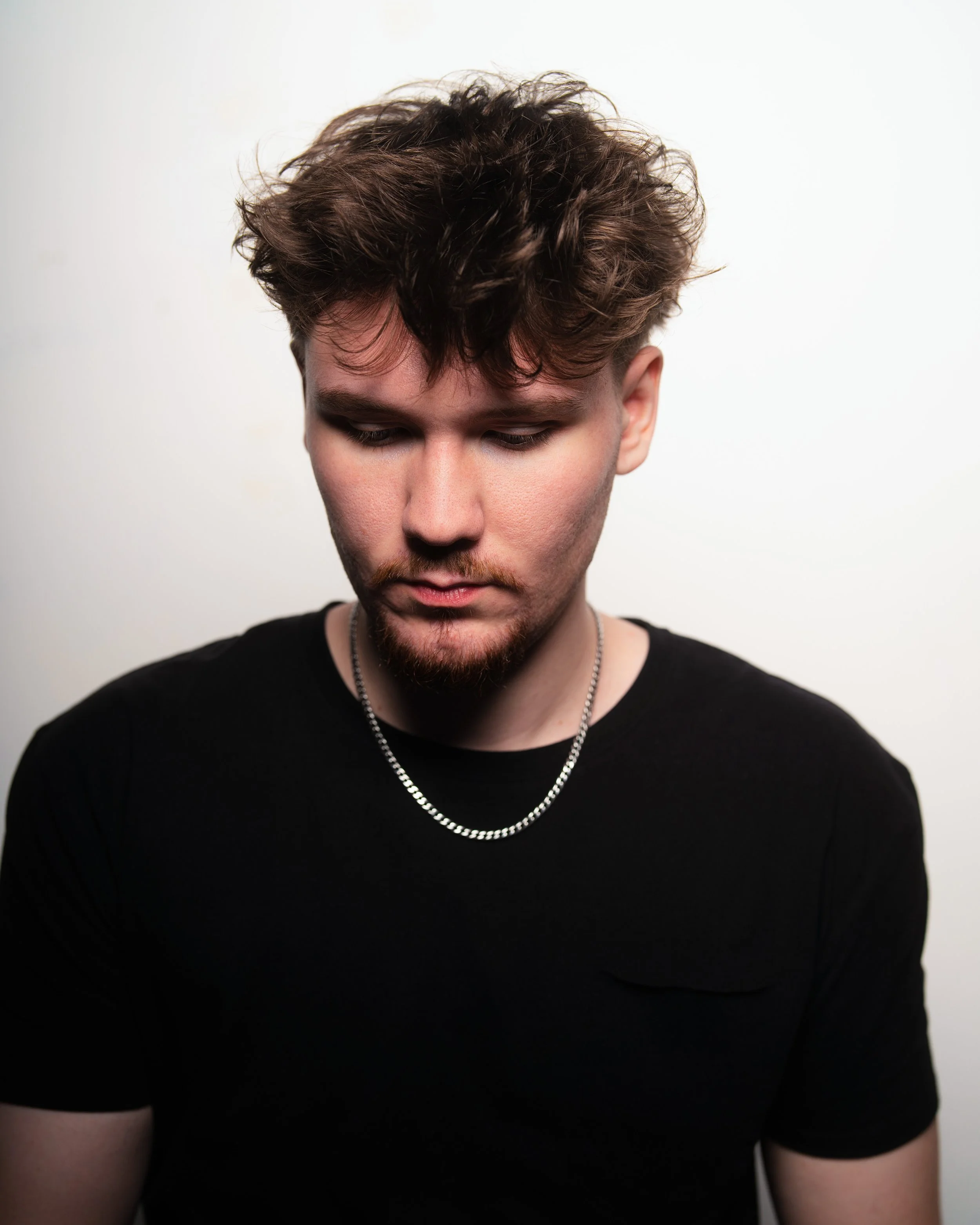 Close-up of a young man with styled, wavy brown hair, wearing a black shirt, silver chain necklace, and looking down against a plain white background.